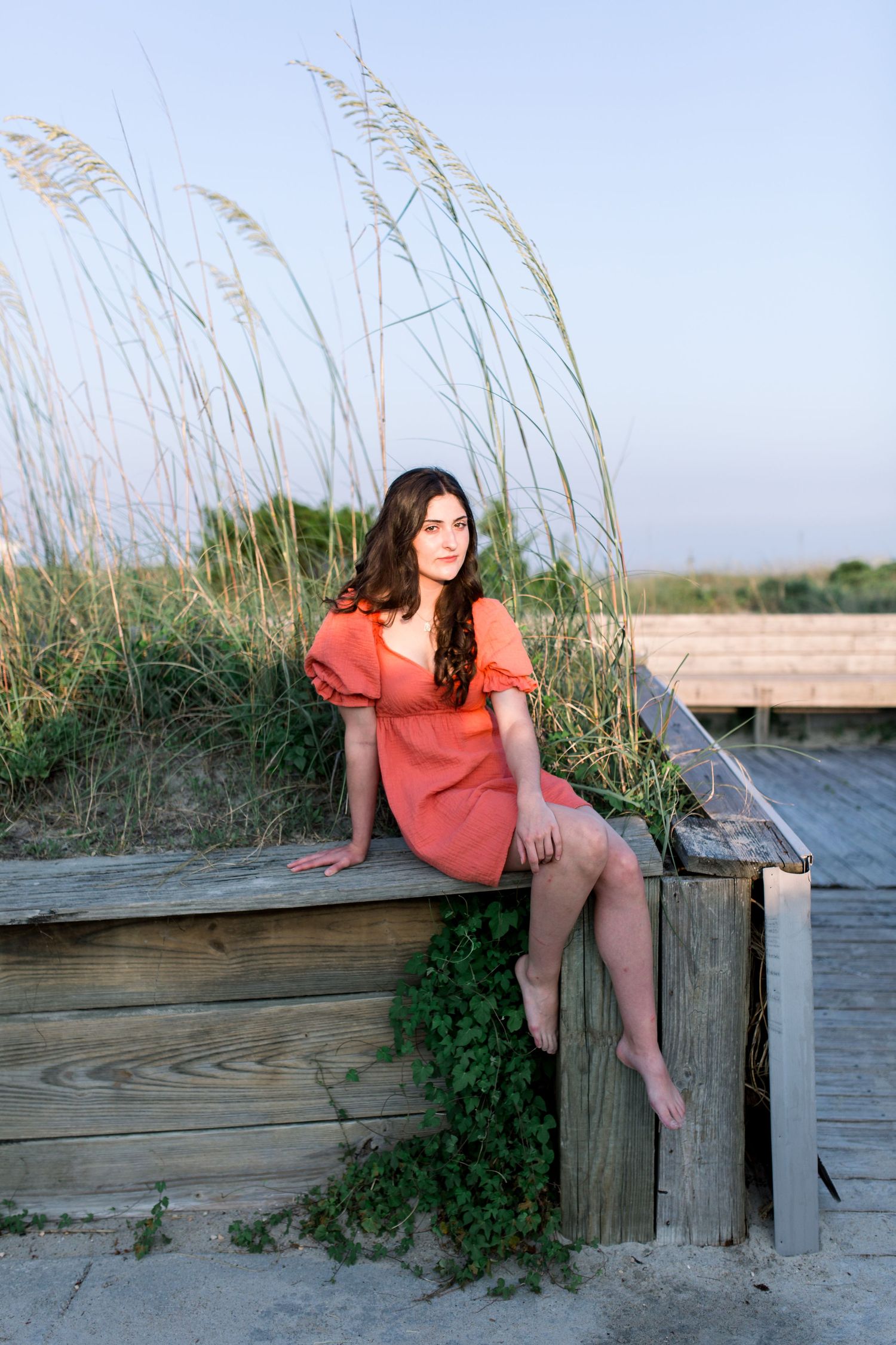 A coral dress-clad figure sits on a wooden boardwalk by the beach with tall grass swaying in the background.