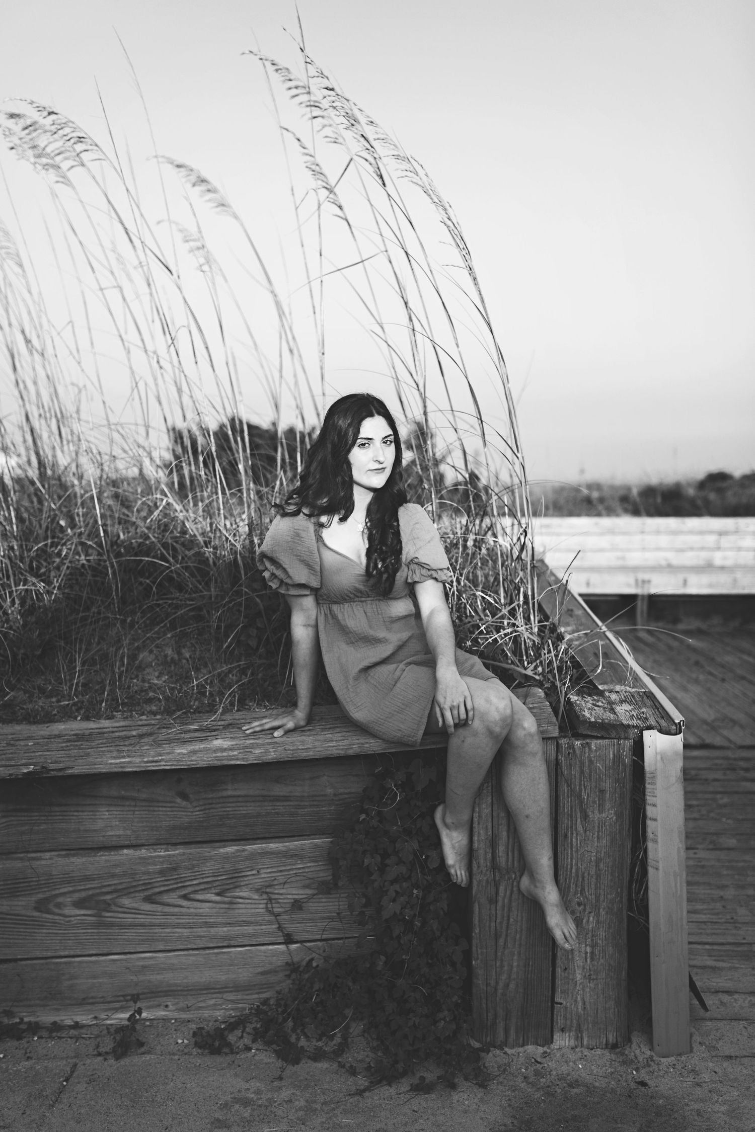 Black and white photo of someone in a dress sitting on a wooden deck with sea grass in the background.