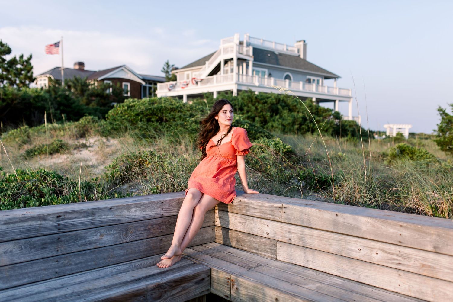 Person in coral dress sitting on wooden steps with coastal houses and American flag in background.