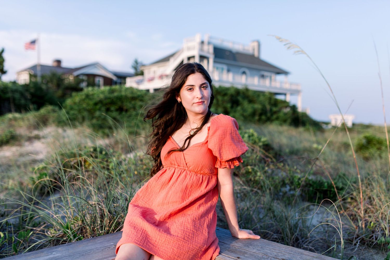 Beachside portrait featuring someone in a coral dress with beach houses and dune grass in background.