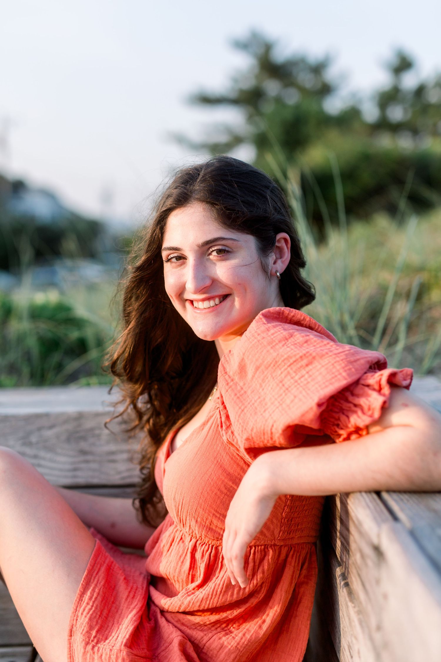 Seaside portrait on wooden deck wearing coral dress with beach grass and coastal scenery.