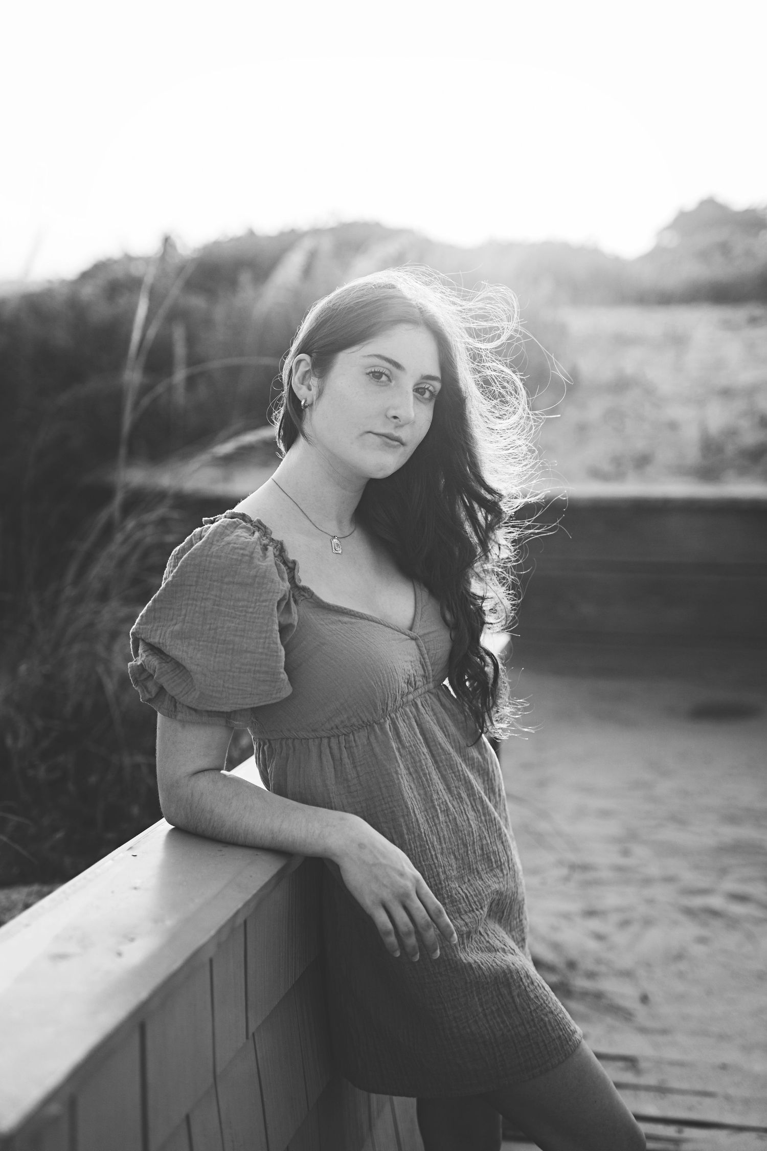 Monochrome portrait with figure in vintage-style dress leaning on railing at golden hour.
