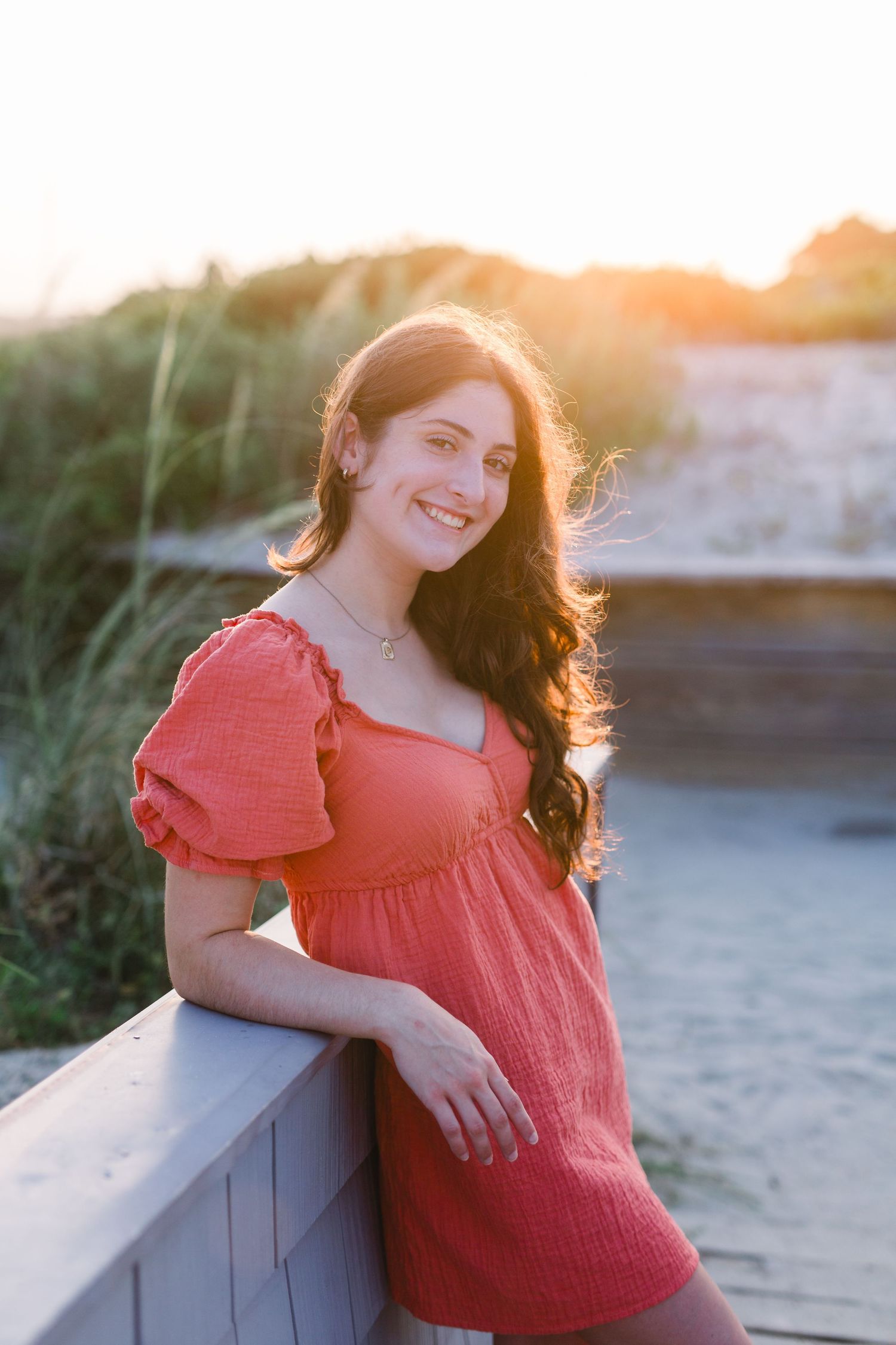 Figure in coral dress poses against railing during sunset with natural backlighting.