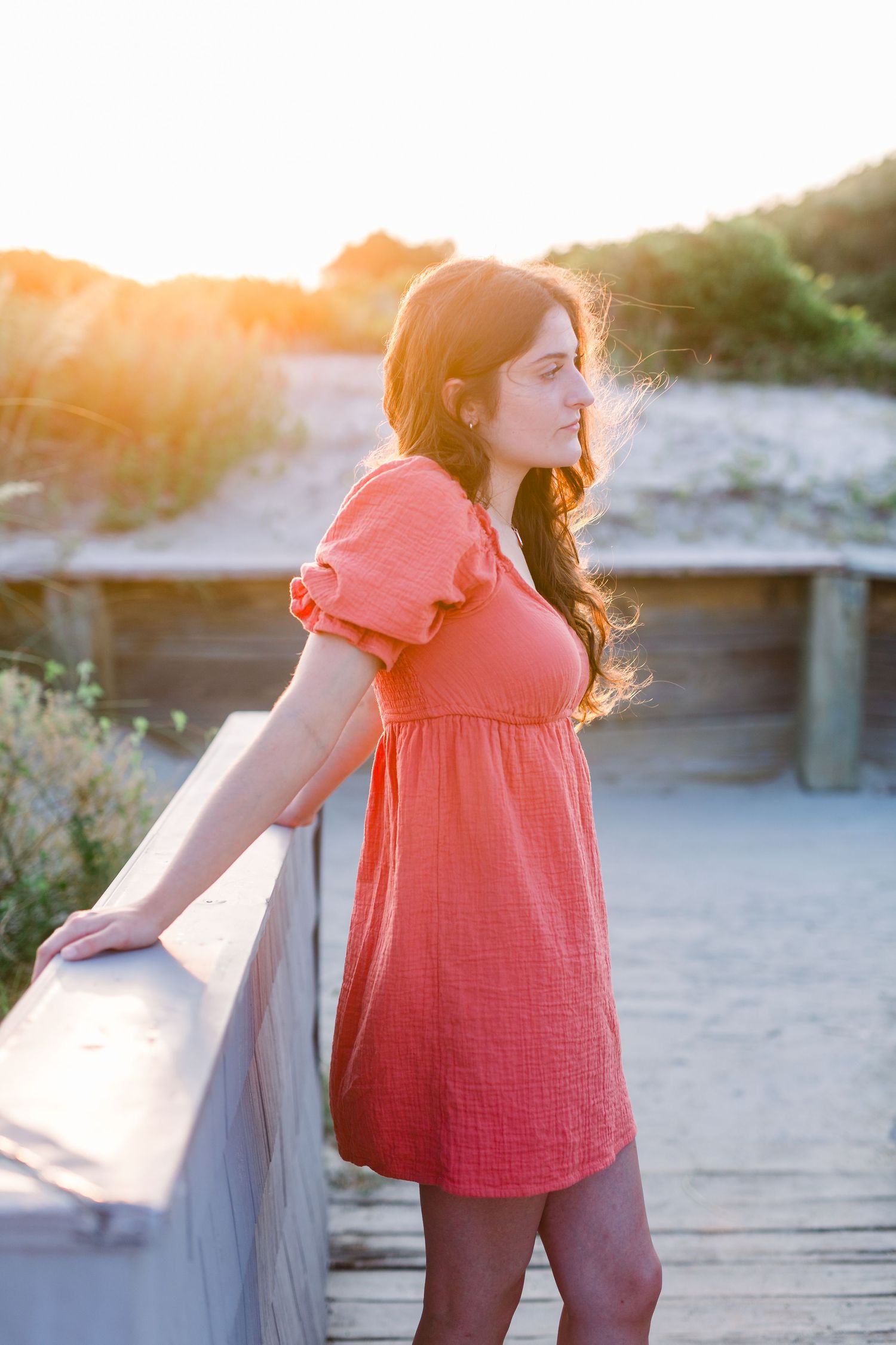 Figure in coral dress leaning on boardwalk railing during golden hour sunset.