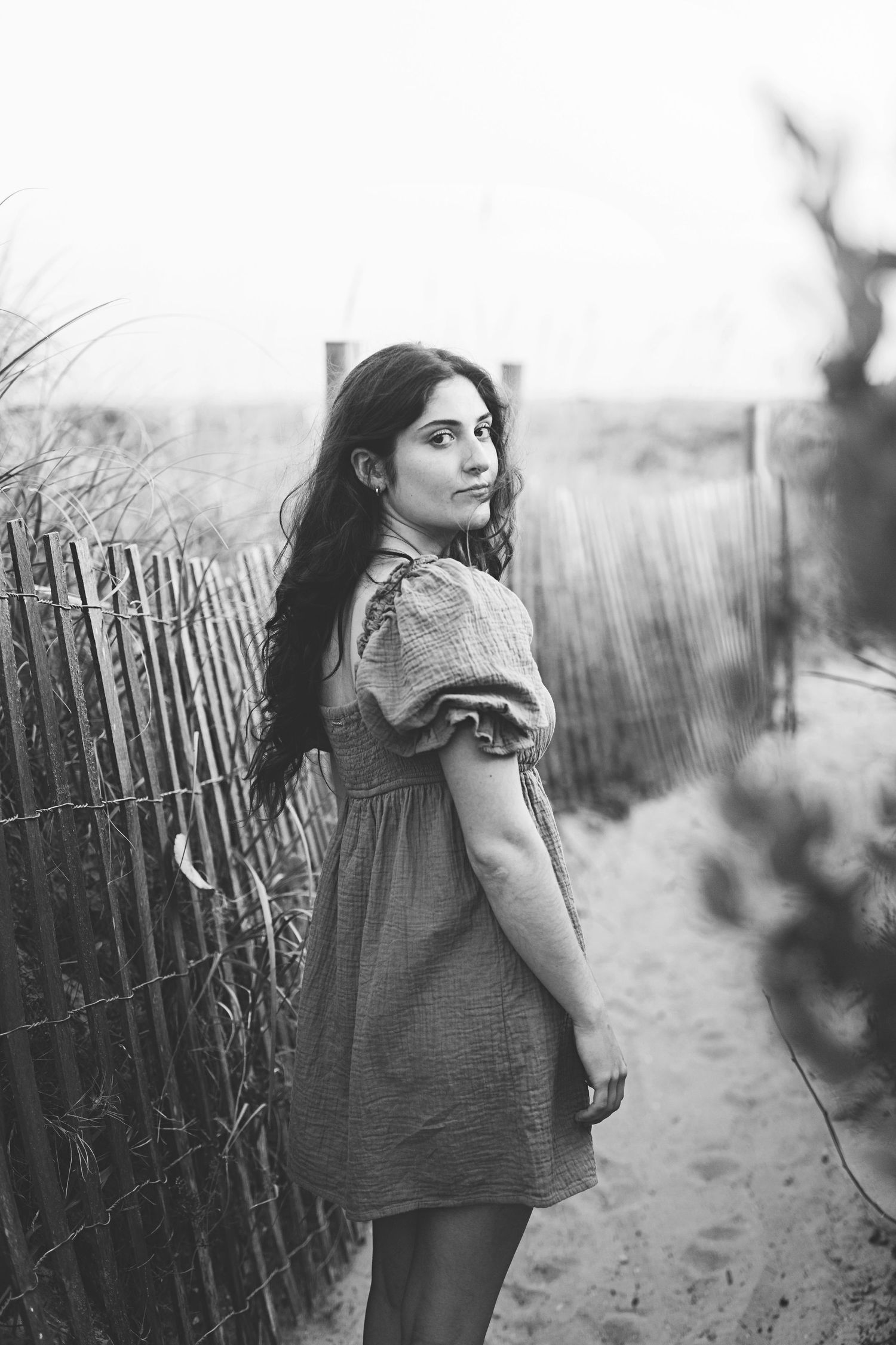Black and white image showing figure in dress posing by wooden fence on beach path.