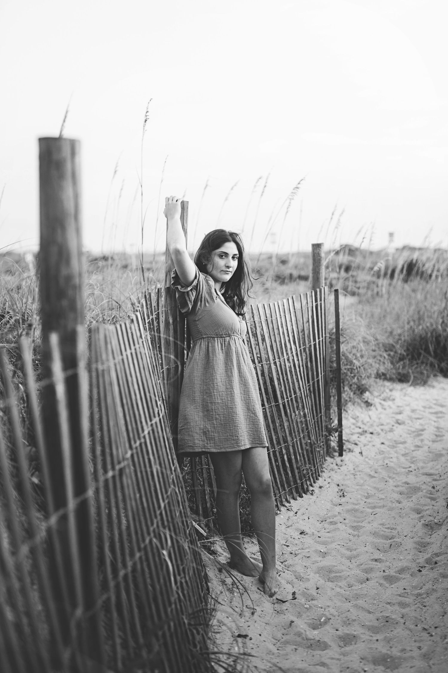 Black and white beach portrait with figure standing by wooden post and sand fence.