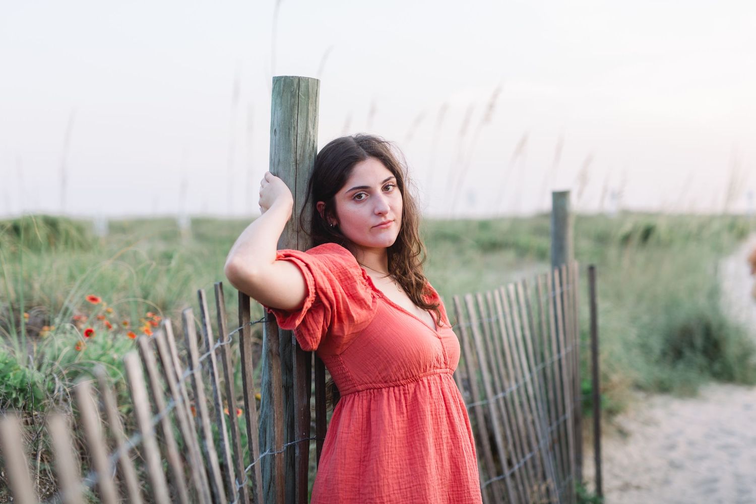 Portrait at beach with figure in coral dress posing against wooden post at dusk.