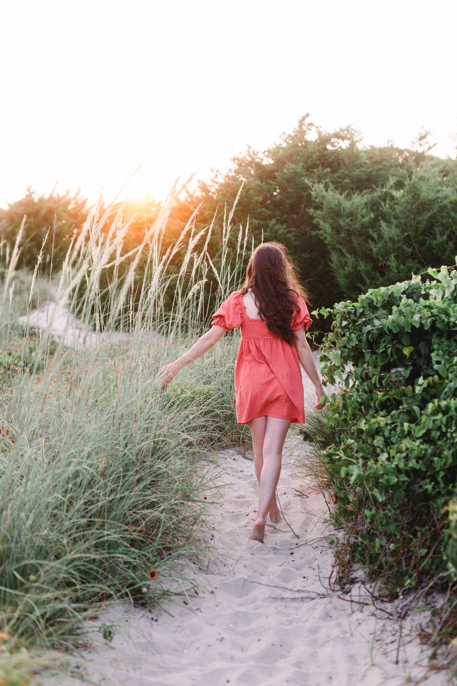 Walking away shot of figure in coral dress on sandy beach path during sunset.