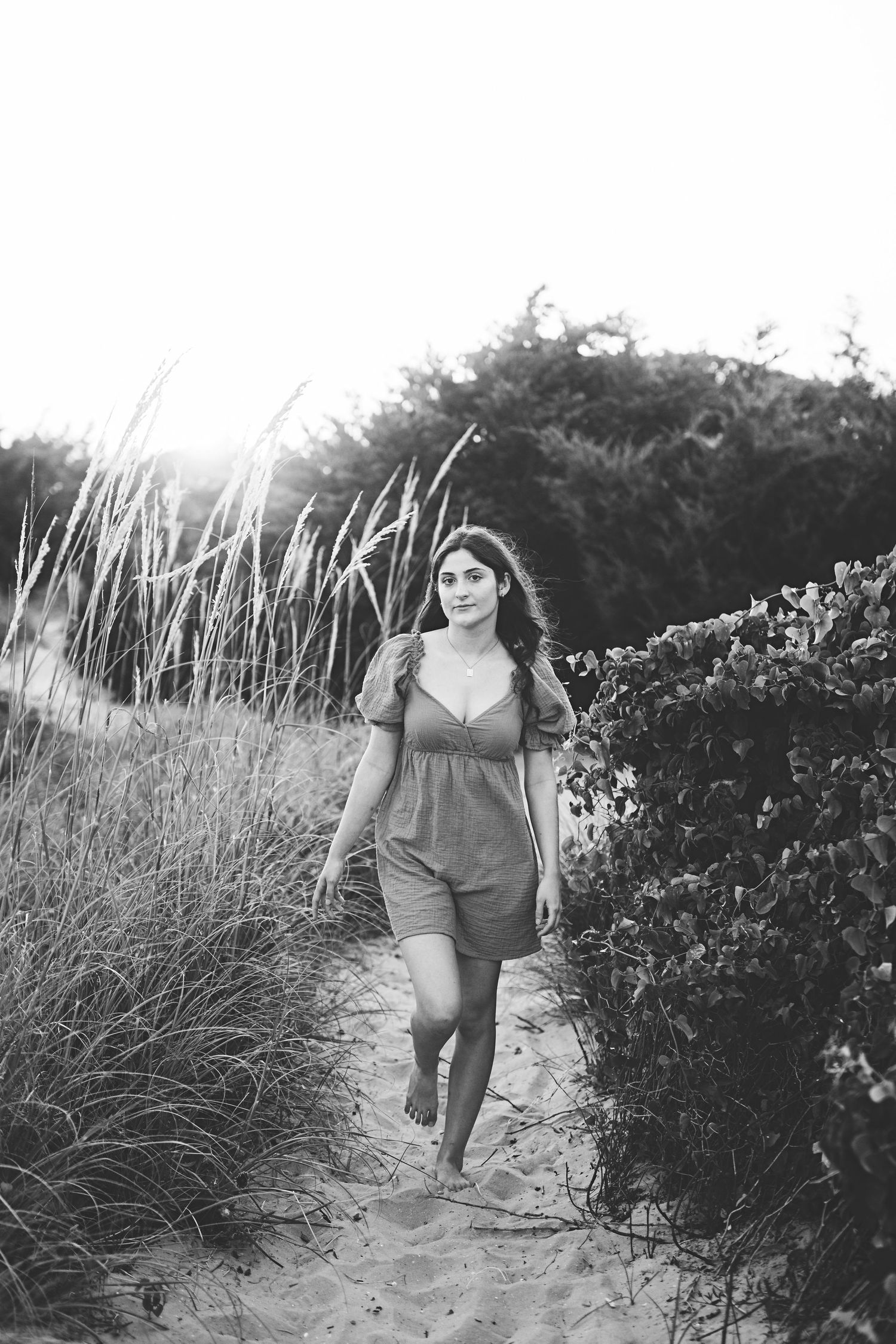 Person walking on sandy beach trail wearing a dark romper surrounded by coastal grasses and shrubs.