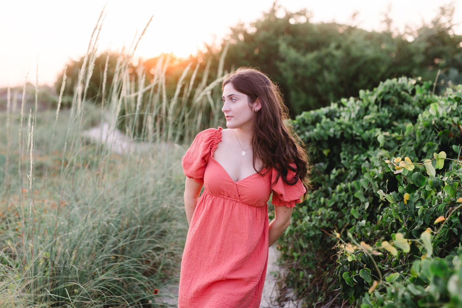 Person in coral dress walking along coastal path with tall grass and greenery at sunset.