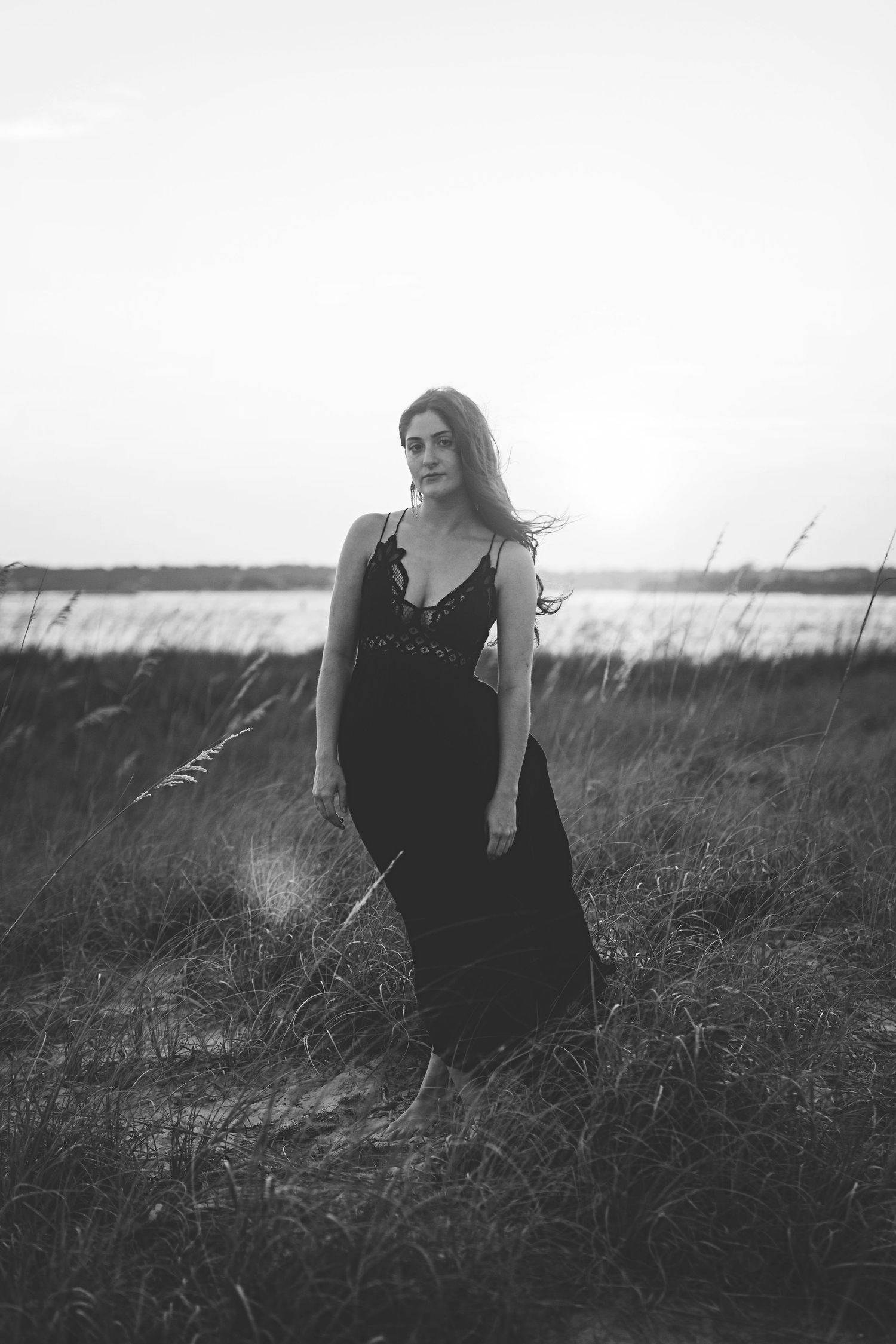 Figure in black dress stands among coastal grasses with water in background in monochrome.