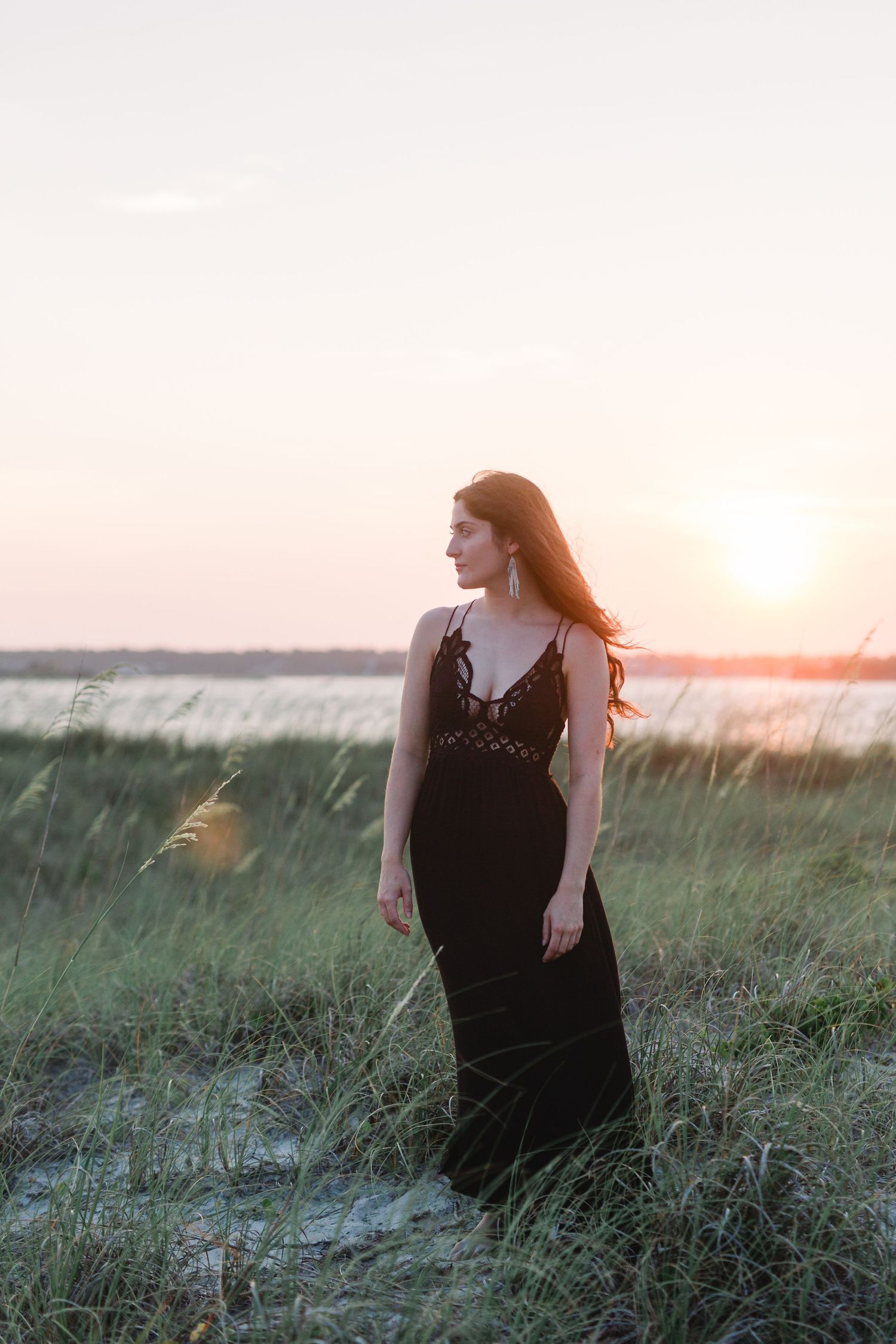Person in black maxi dress stands in coastal field at sunset with water in background.