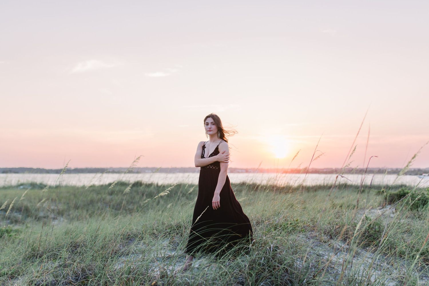 A figure in a flowing black dress poses gracefully in a grassy field at sunset.