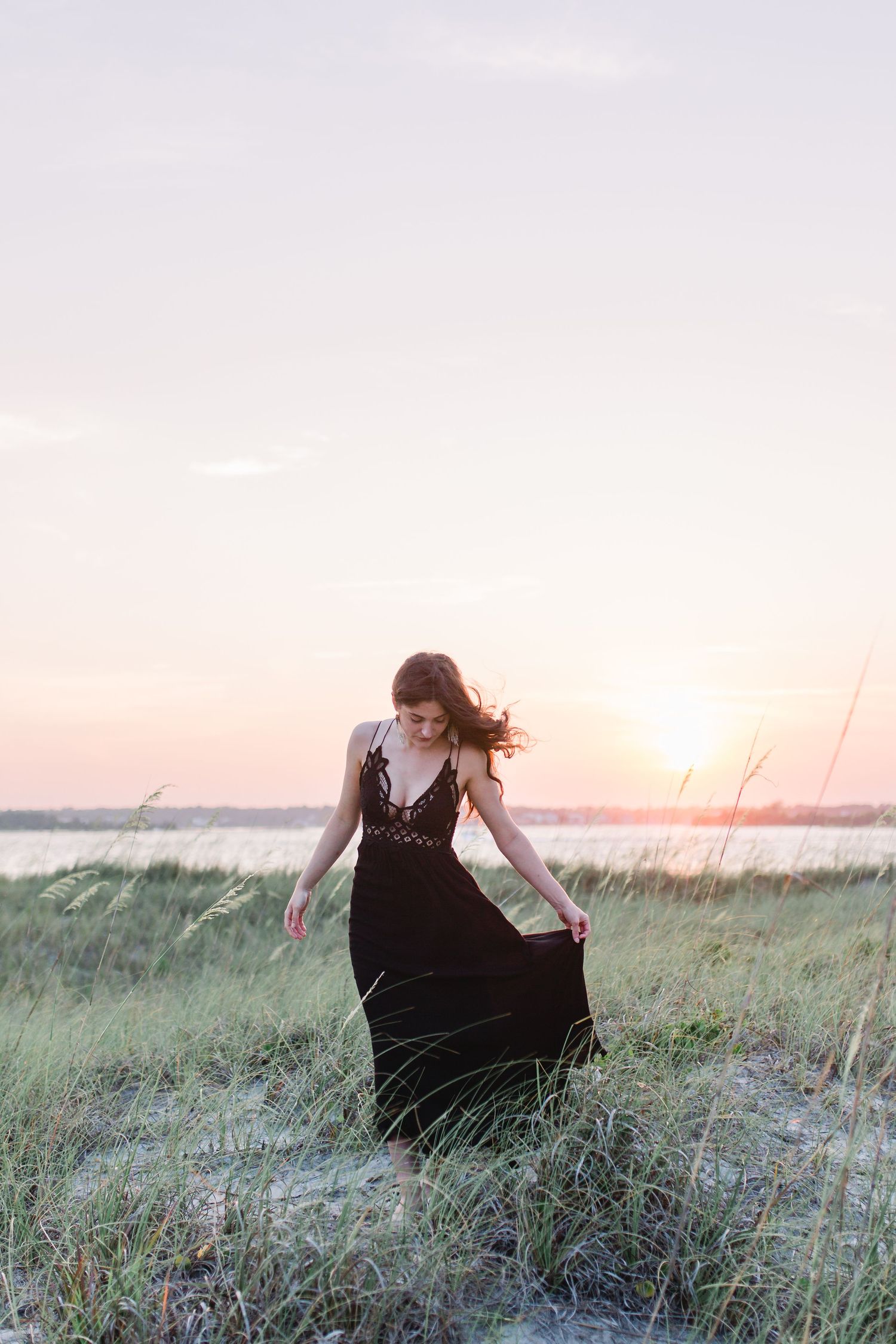 A black dress twirls against a sunset backdrop in tall coastal grass.