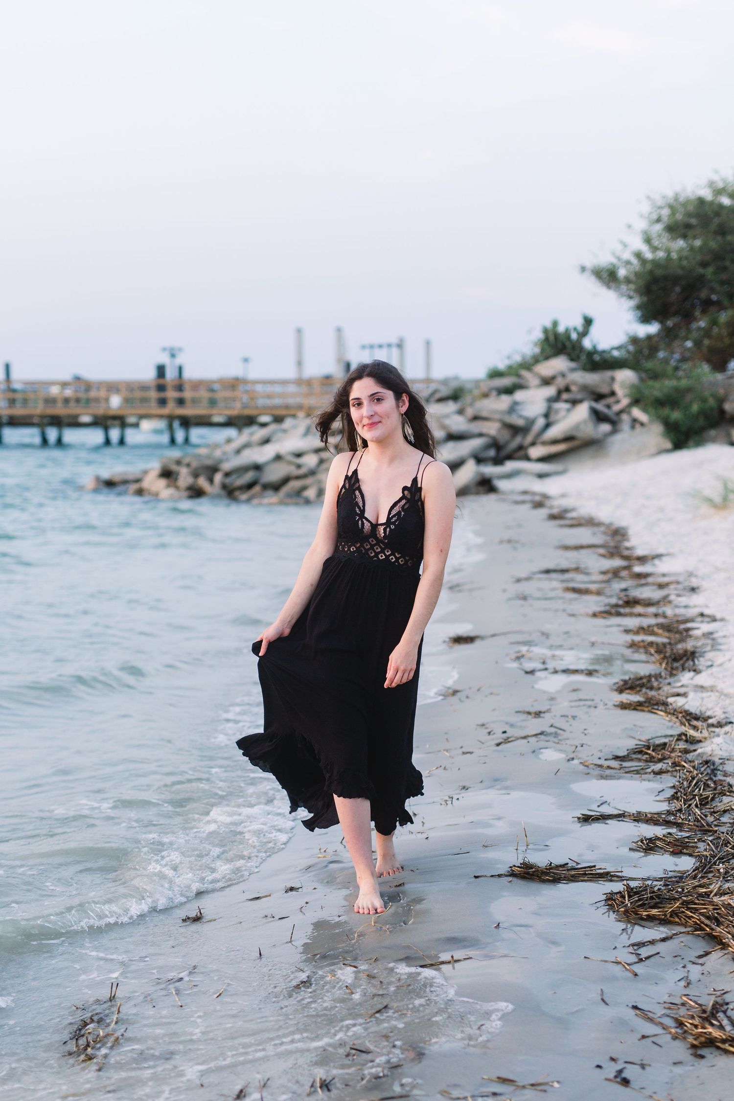 Person in black dress walking barefoot along a rocky beach shoreline.