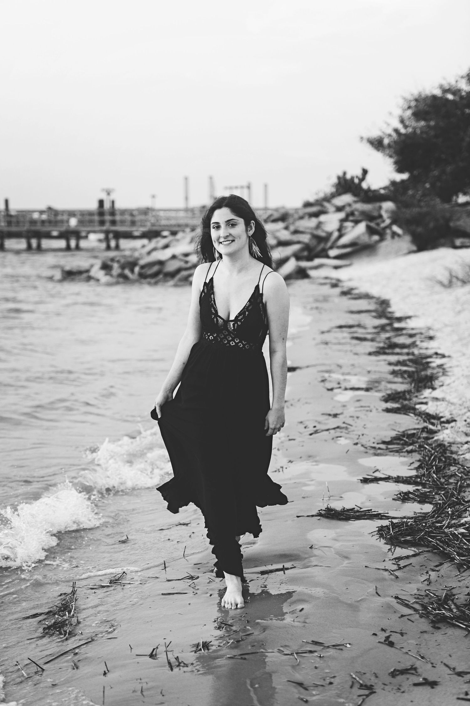 Black and white photo of someone walking along a wave-washed beach near a pier.
