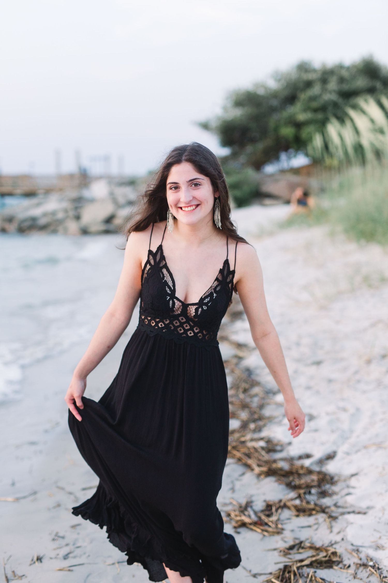 Someone in a flowing black dress smiles while walking on a coastal beach.