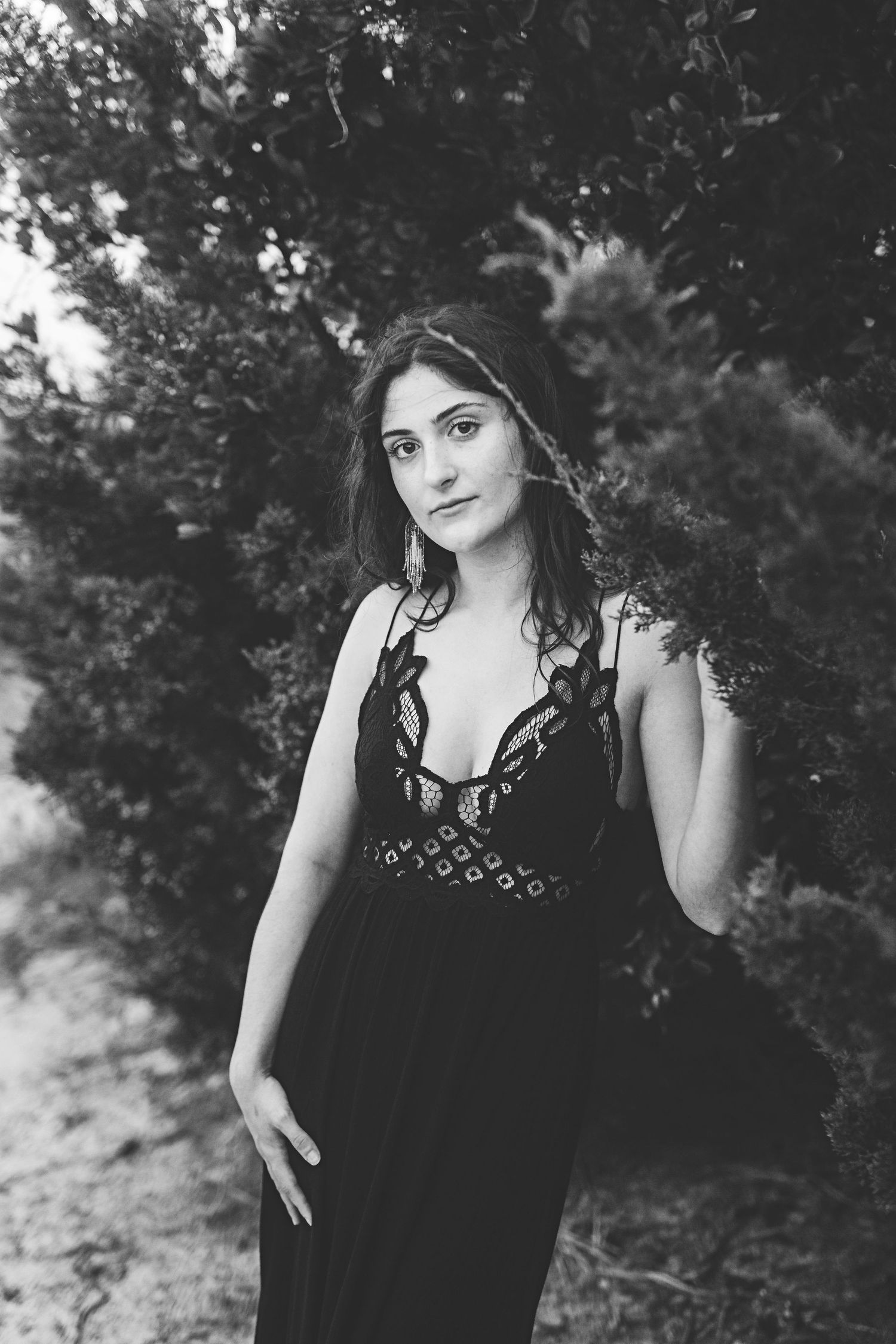 A black and white portrait of someone in an elegant black lace dress with natural foliage in the background.
