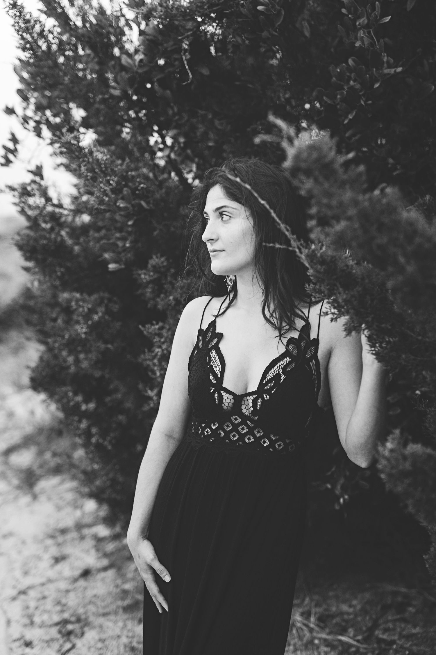 A monochrome fashion photo showing a person in a dark formal dress posing against evergreen trees.