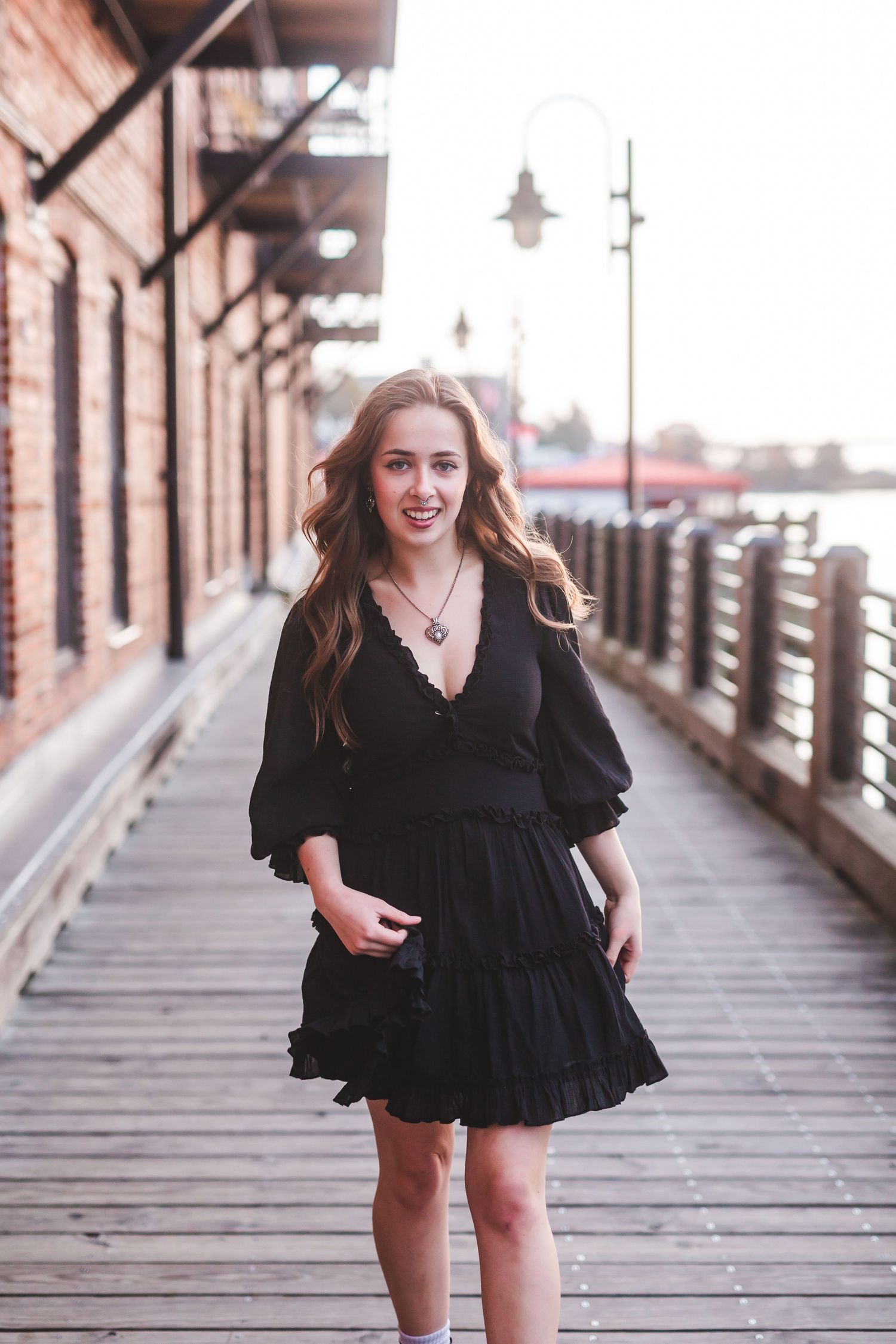 Woman in black ruffle dress standing on rustic wooden boardwalk near brick building.