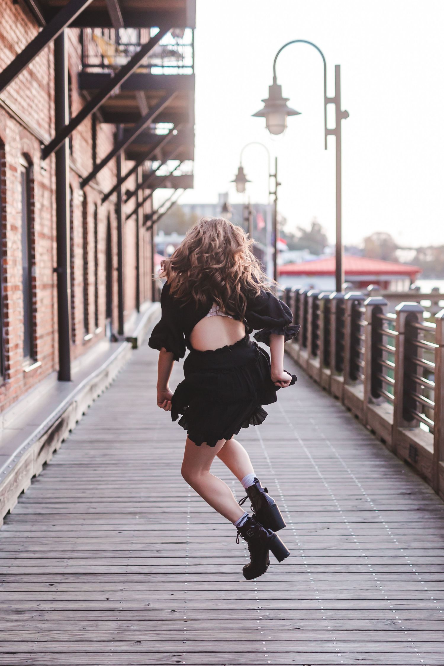 Person in black dress jumping playfully on wooden boardwalk with brick building and lamp posts.