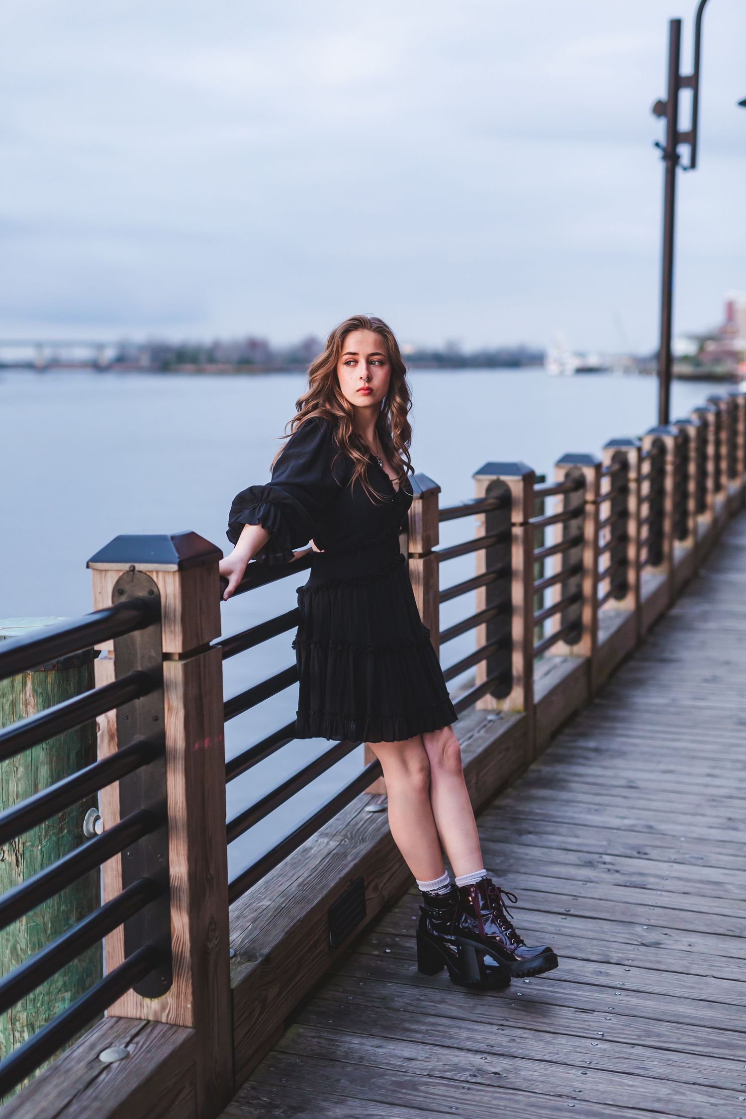 Woman in black dress posing against waterfront railing during overcast day.