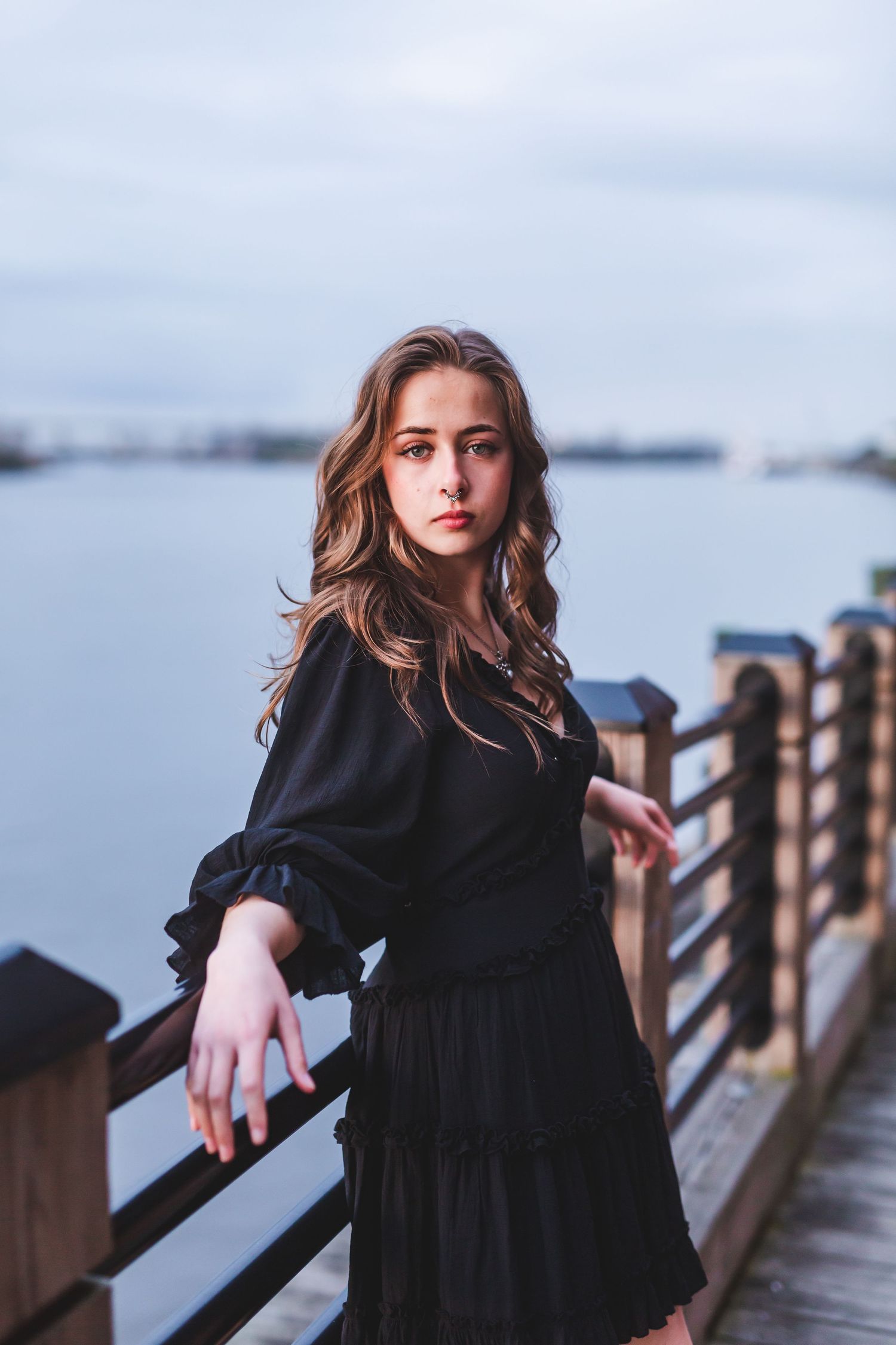 Woman in black dress leaning on wooden railing with water and cloudy sky in background.