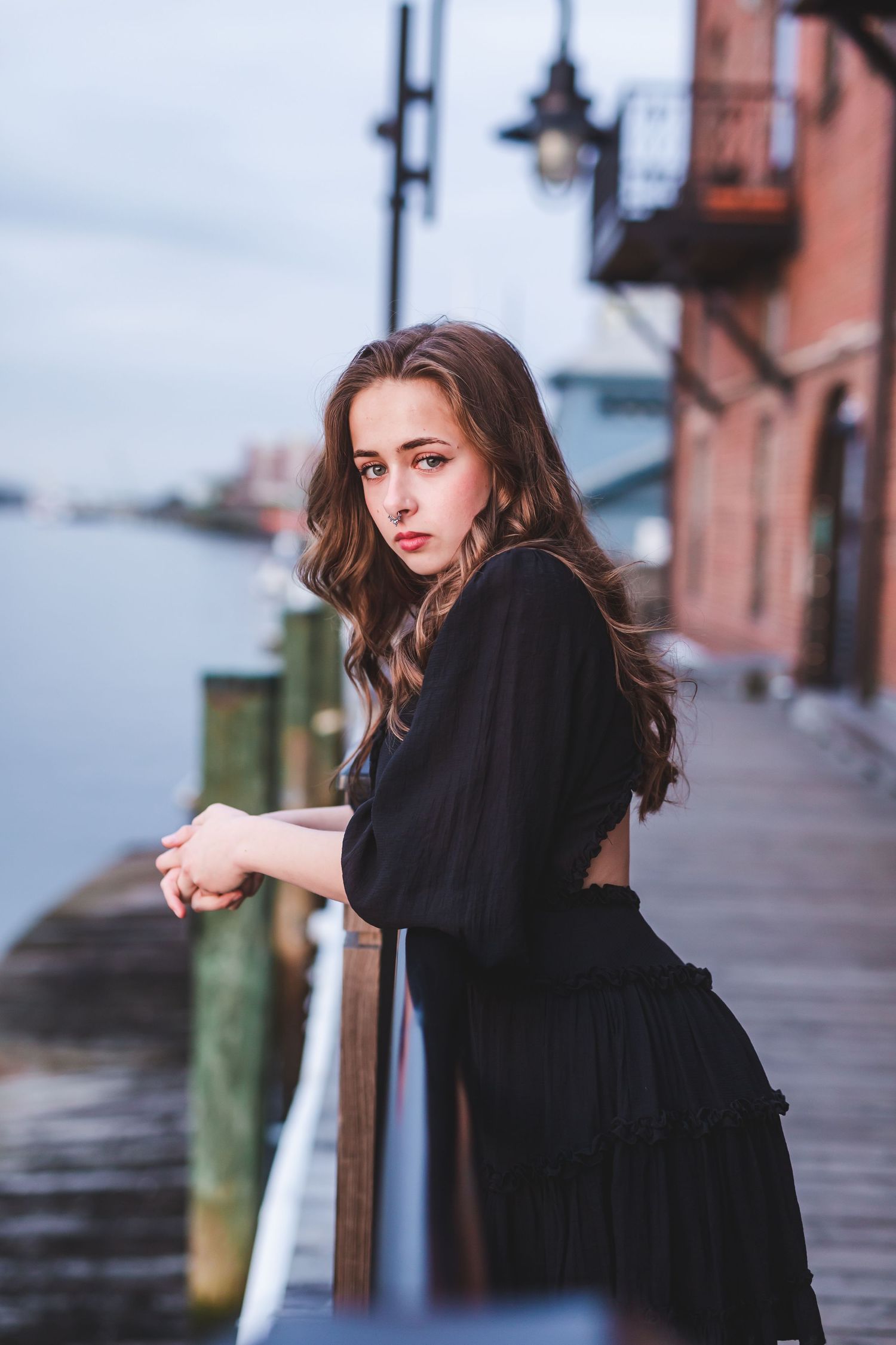 Woman in black dress poses against railing on waterfront boardwalk.