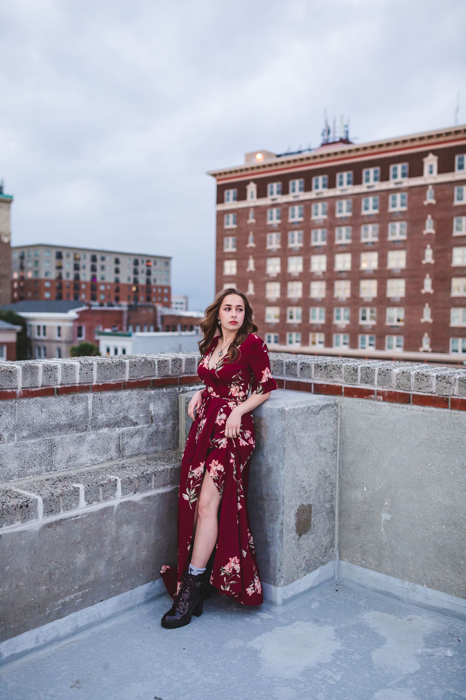 Woman in burgundy floral maxi dress standing on urban rooftop with brick buildings in background.