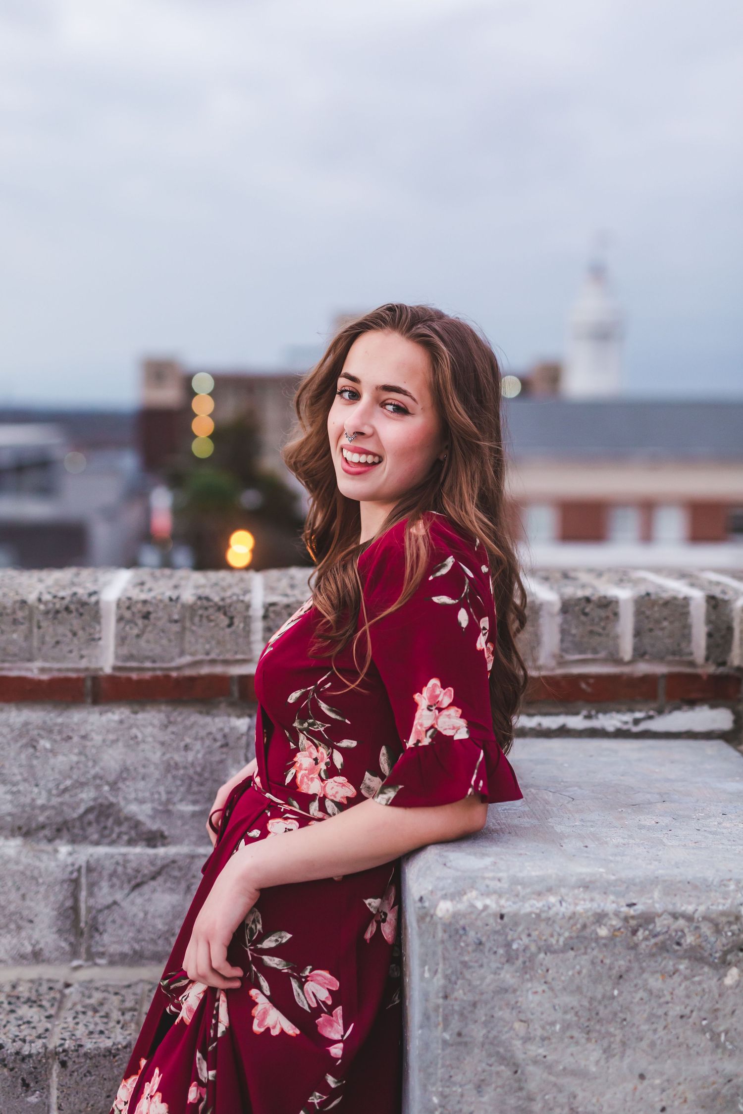 Woman in red floral dress leaning against stone wall with cityscape behind.