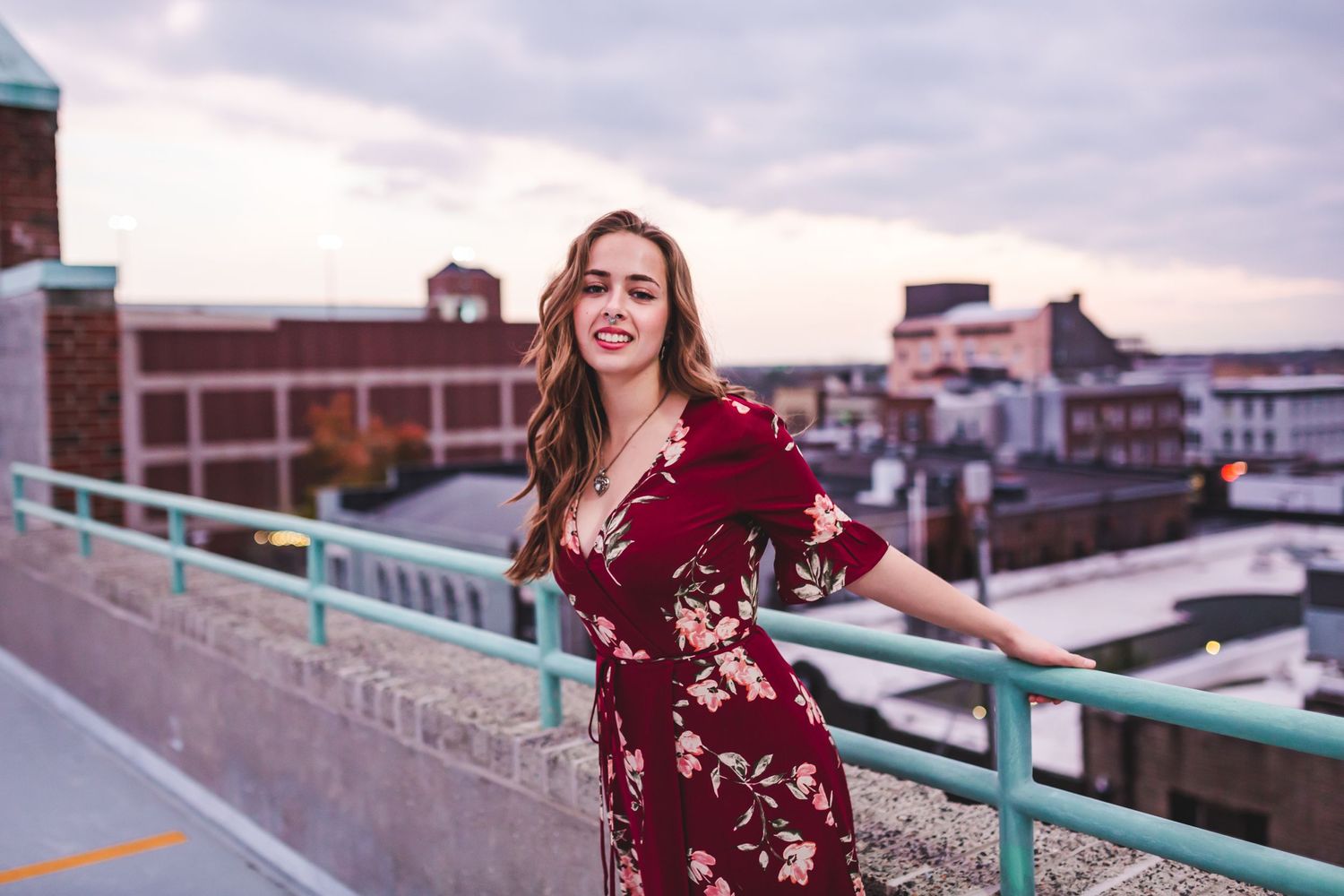 Woman in red floral dress poses on rooftop with city buildings in background.