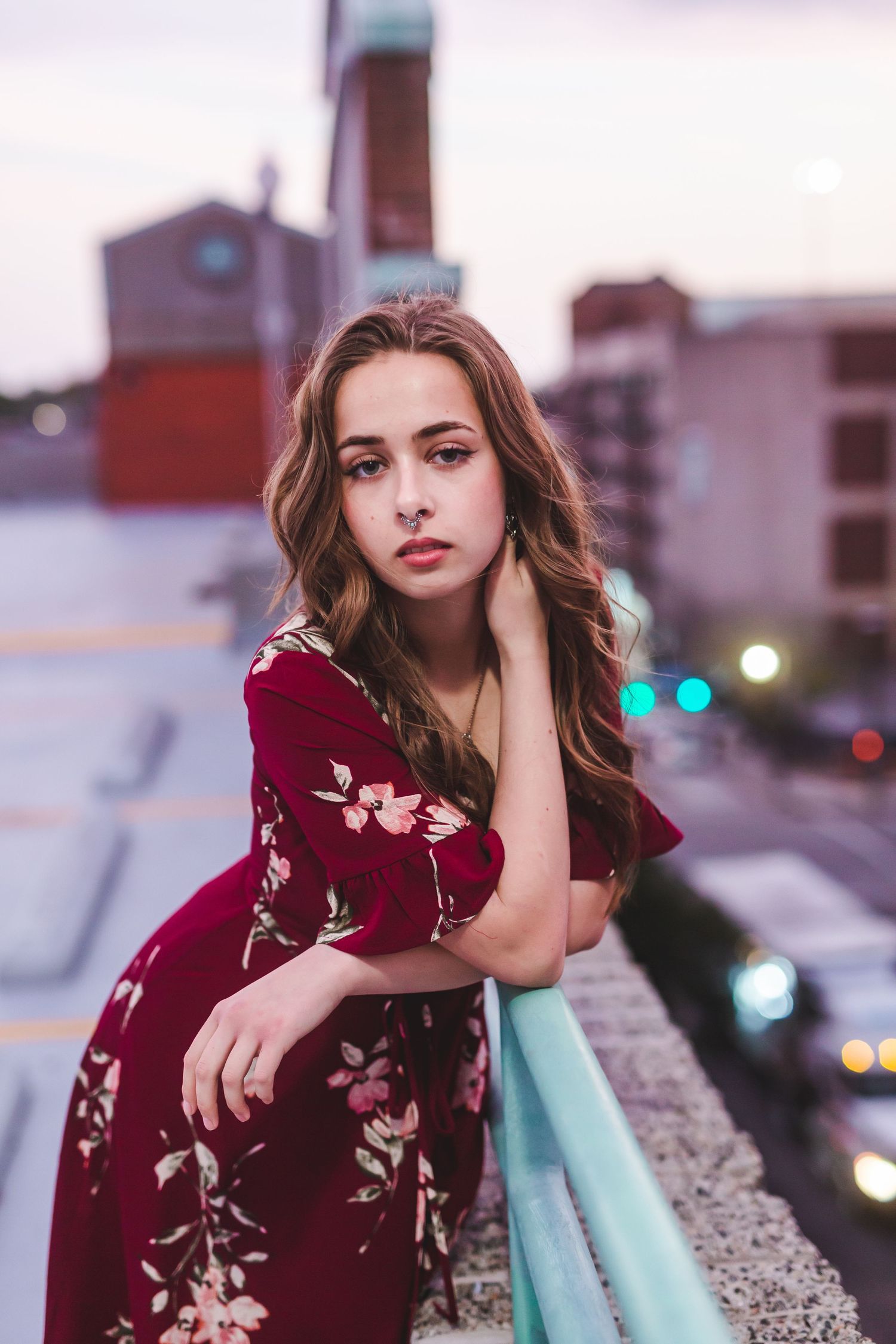 Model in a burgundy floral dress poses against city rooftop railings at dusk.