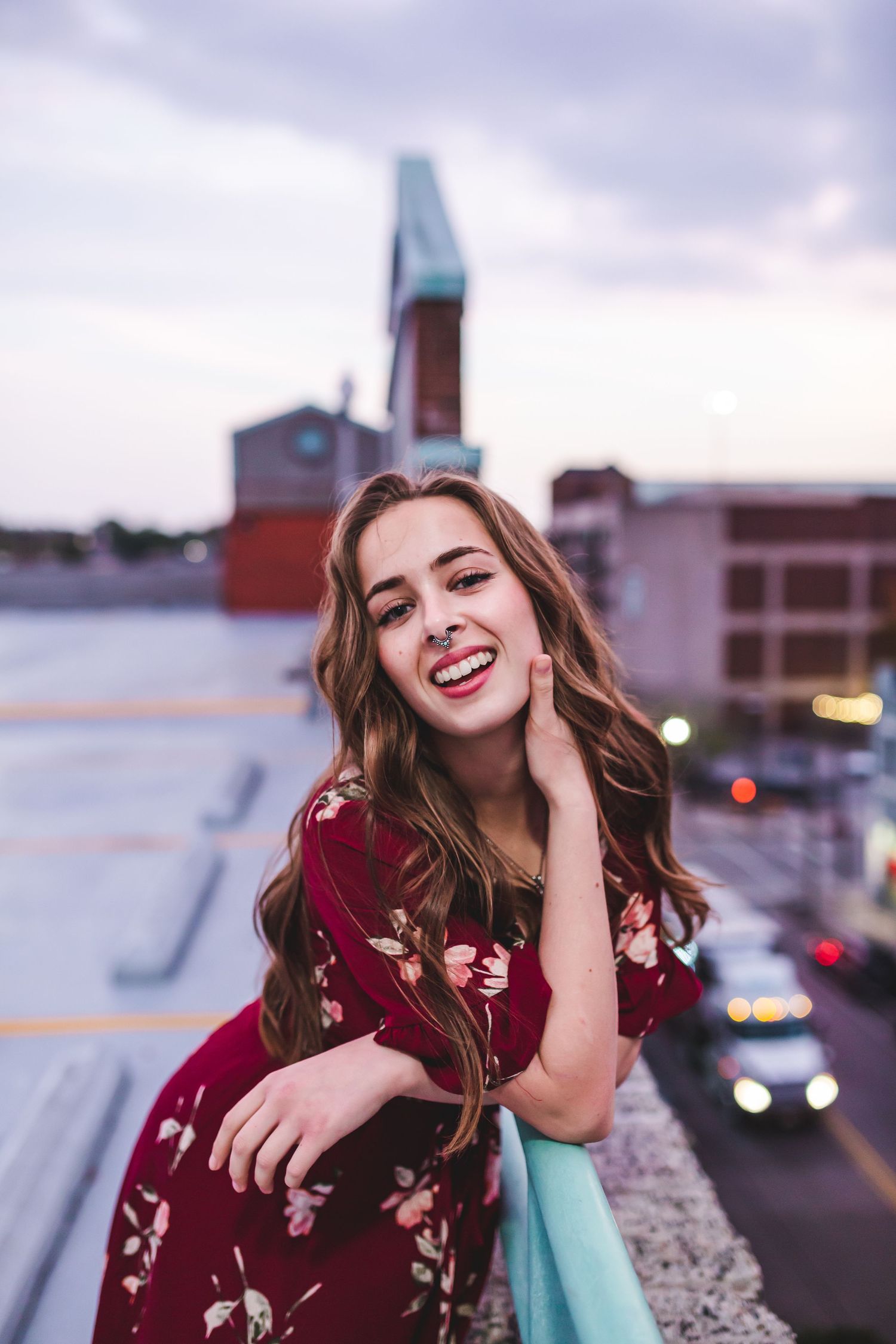 Woman in burgundy dress laughing while leaning on turquoise railing with cityscape behind.
