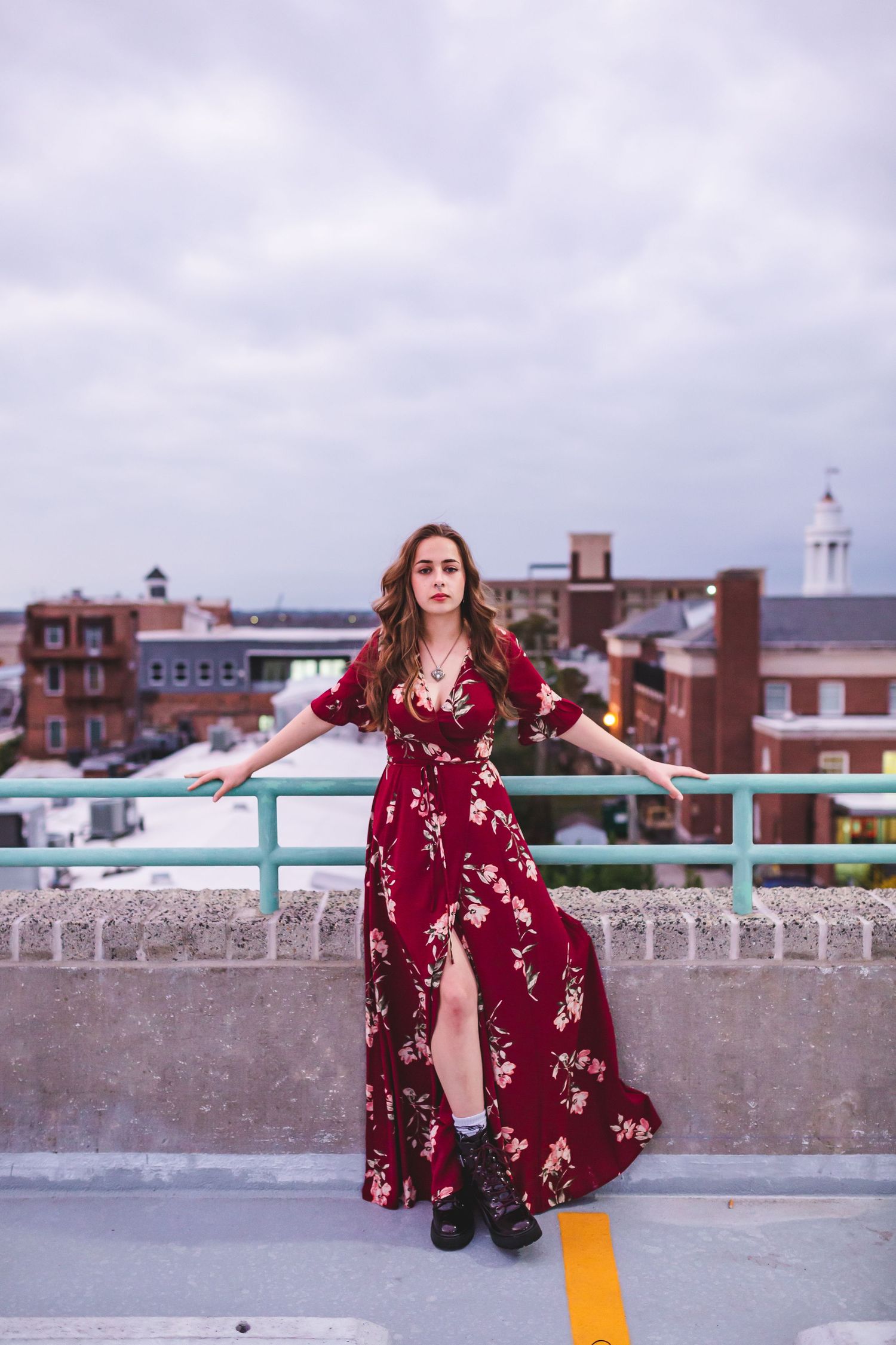 Elegant pose in burgundy maxi dress on urban rooftop with cloudy sky backdrop.