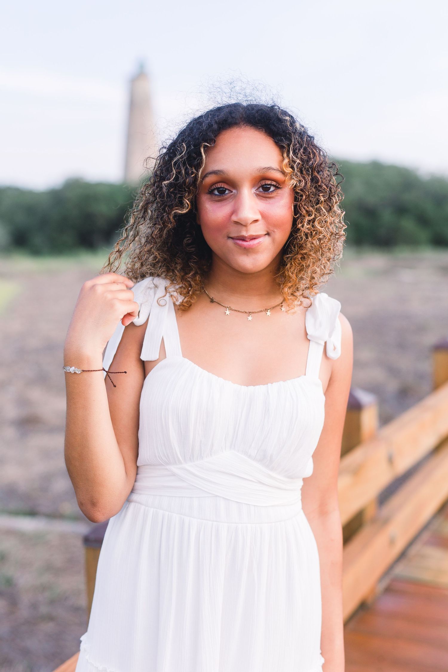 A portrait of someone wearing a white summer dress with bow straps near a coastal boardwalk.