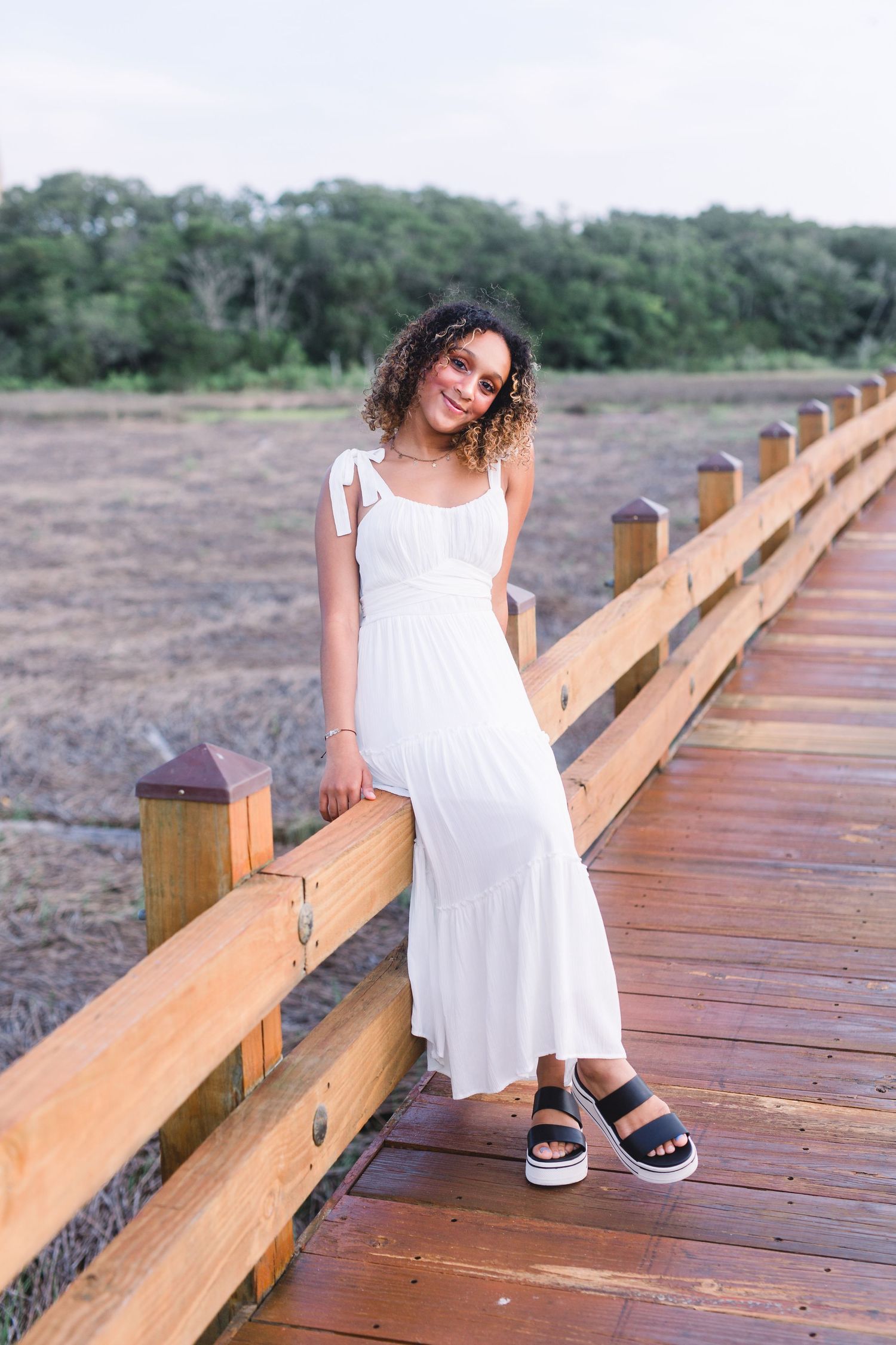 A casual summer portrait on a wooden boardwalk featuring a white maxi dress and platform sandals.