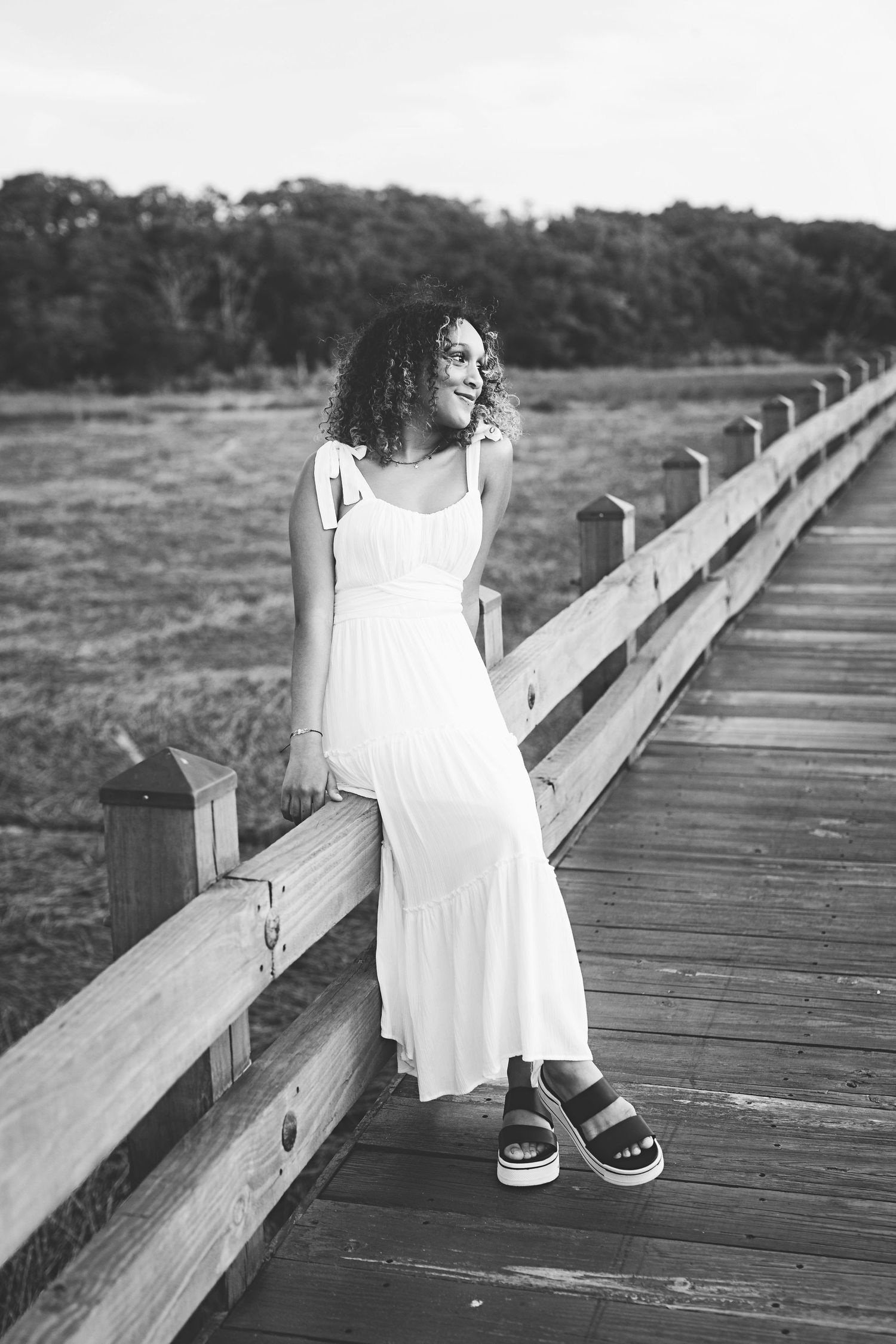 A black and white image of someone sitting on a wooden boardwalk railing in a flowing dress.
