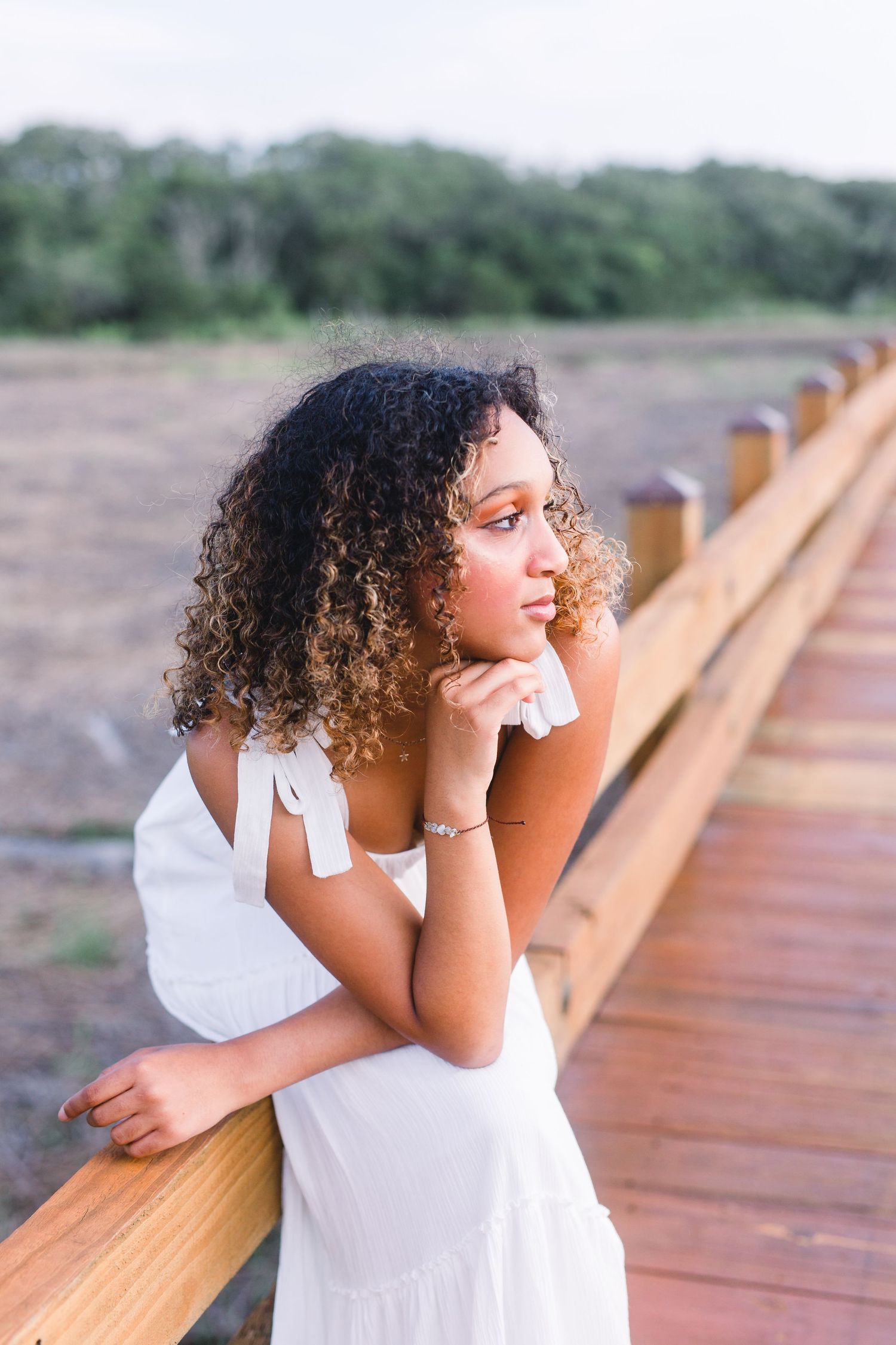 A portrait of someone in a white dress leaning thoughtfully on a wooden boardwalk railing.