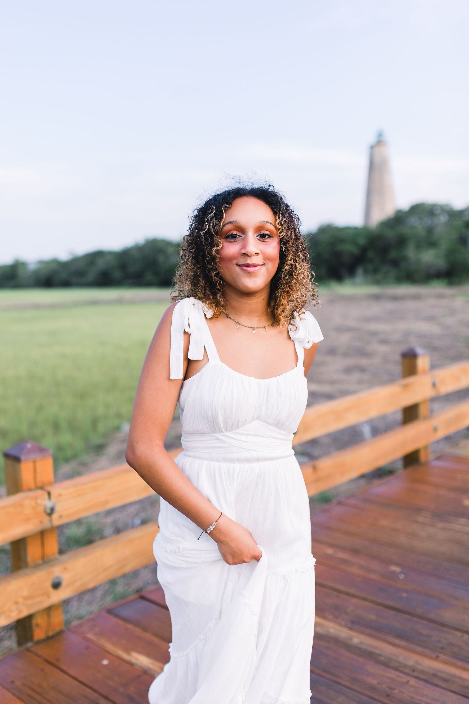 Woman in white sundress poses on wooden boardwalk with lighthouse visible in background during summer day.