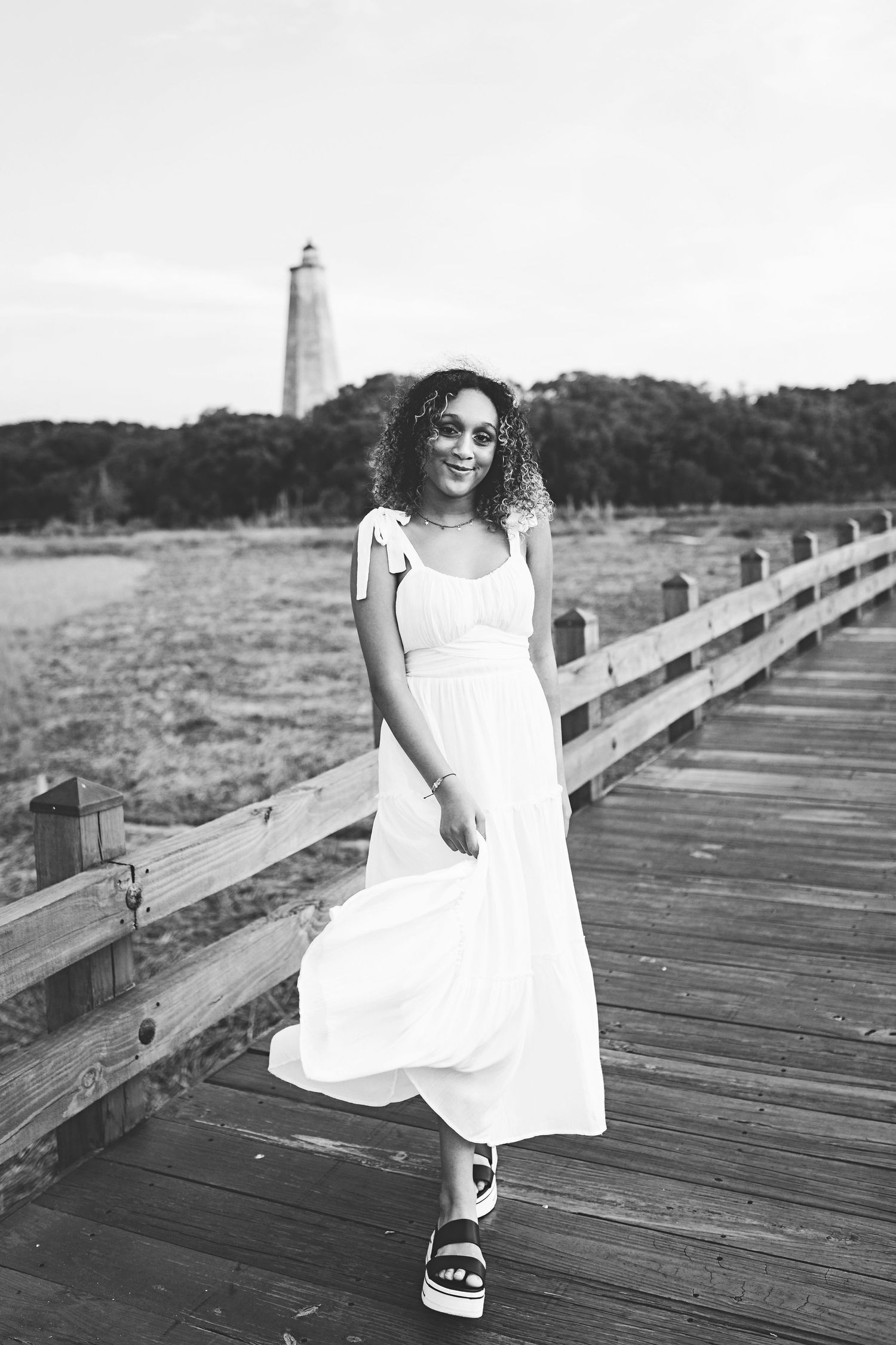 Black and white portrait of woman in flowing white dress and sandals on wooden boardwalk near lighthouse.