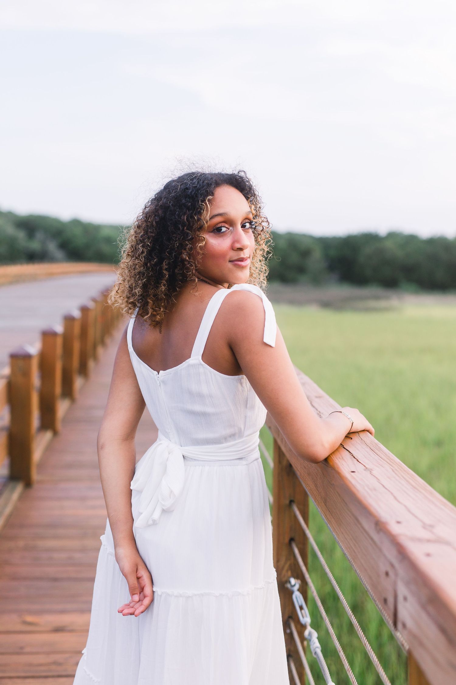 Woman in white tie-strap dress leans against railing of wooden boardwalk overlooking grassy field.