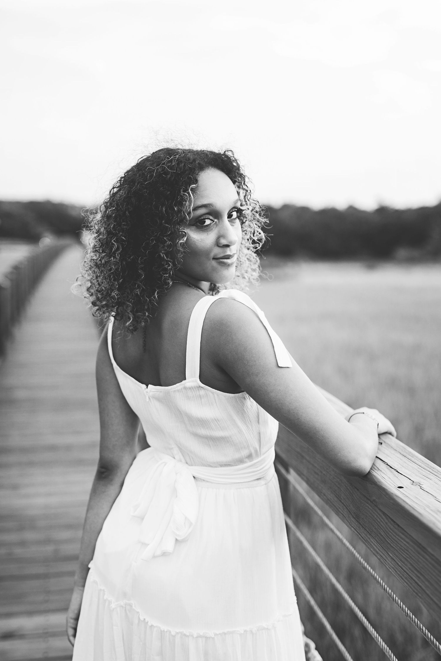 Black and white photo of woman in white dress looking over shoulder while standing on wooden boardwalk.