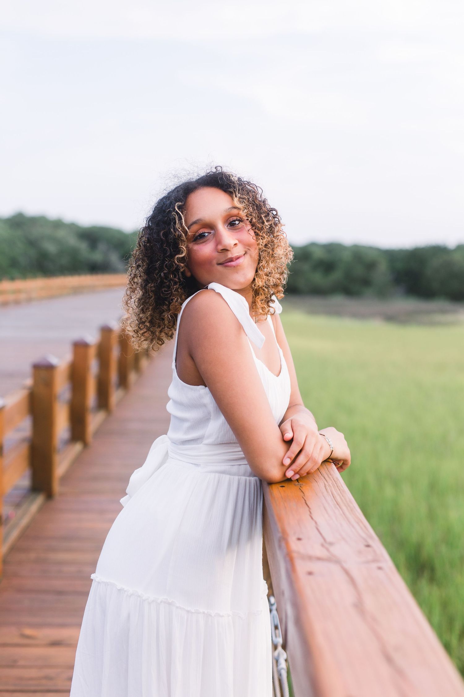 Woman in flowing white summer dress poses against wooden railing with scenic grassland backdrop.