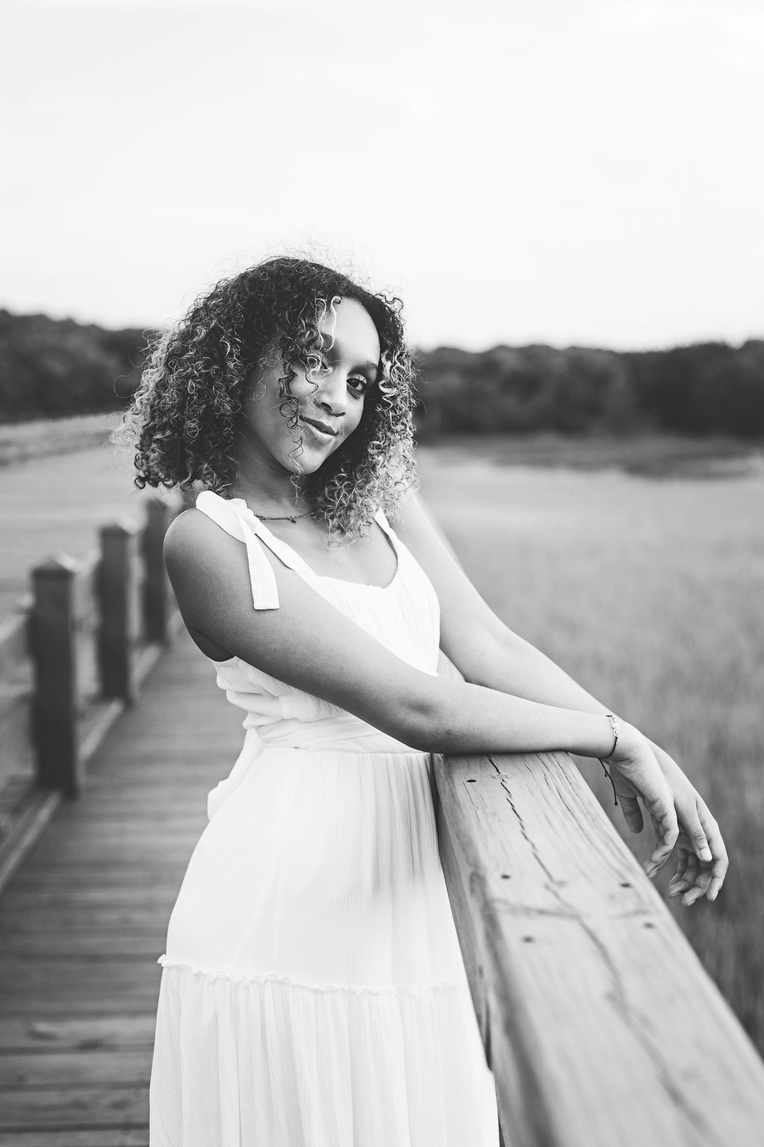 Black and white portrait of woman in white dress leaning on wooden railing during outdoor photoshoot.