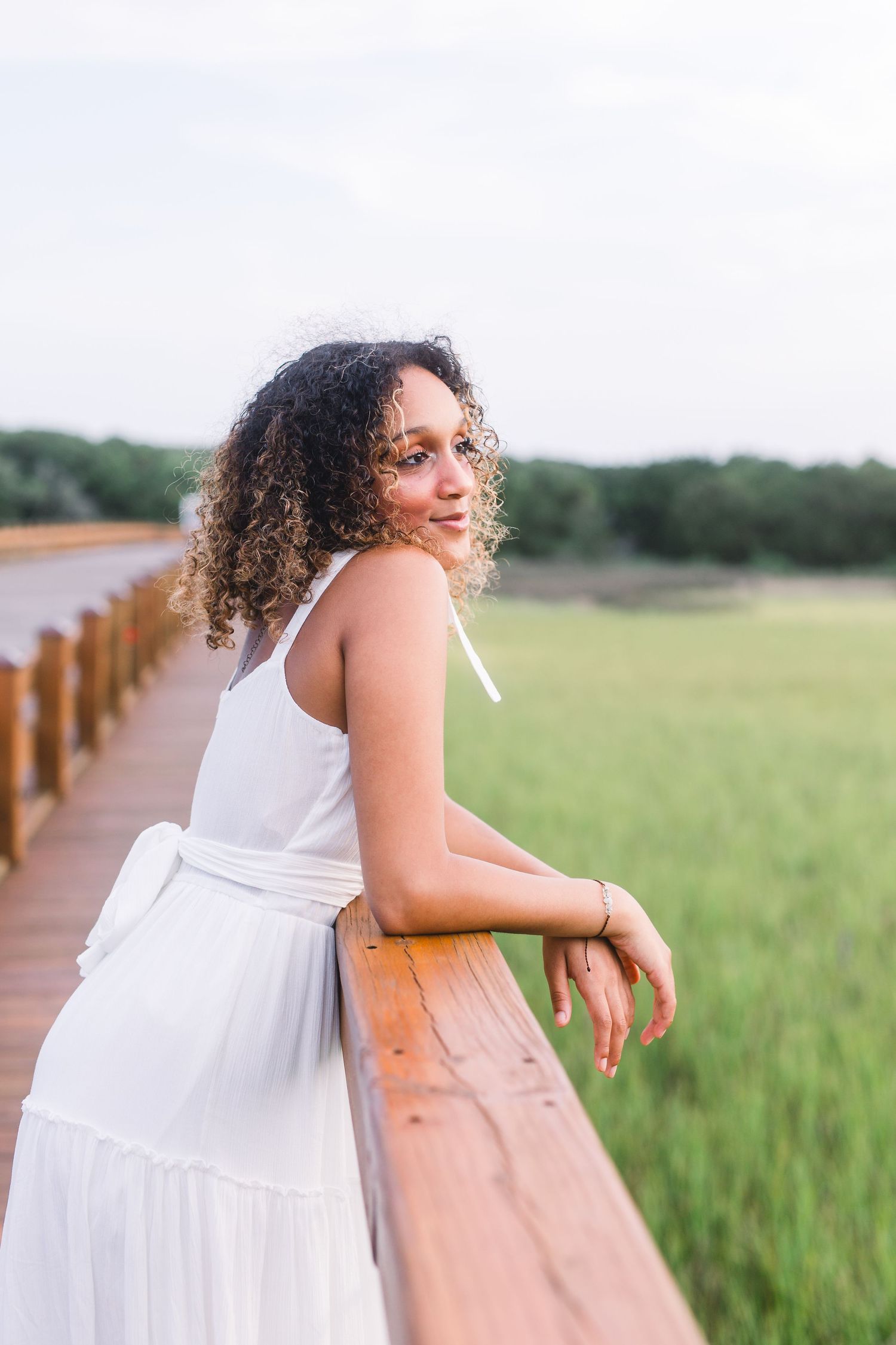 Woman in white tiered dress leans casually against wooden railing of coastal boardwalk.