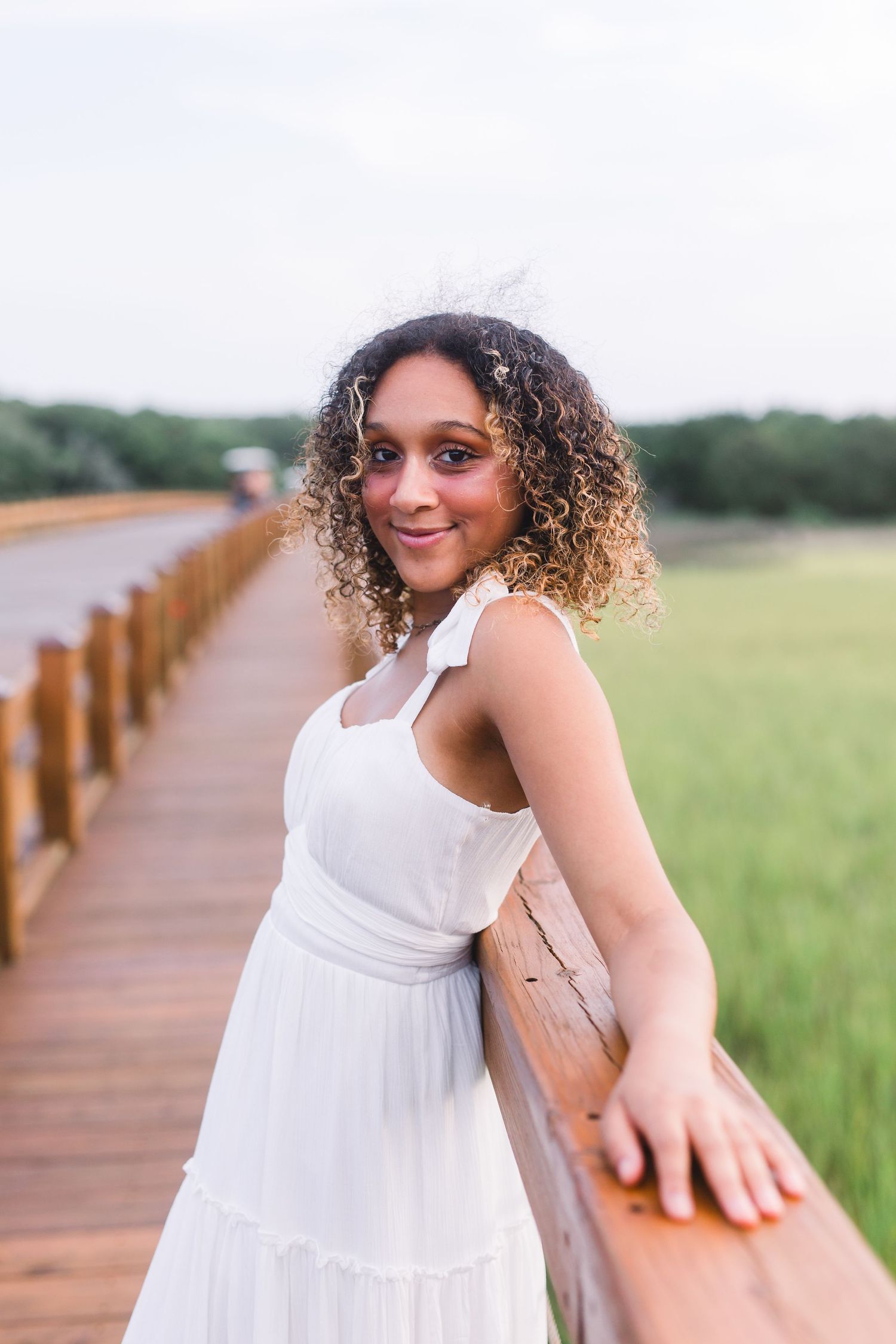 Woman wearing white summer dress poses playfully on wooden boardwalk with marsh view.