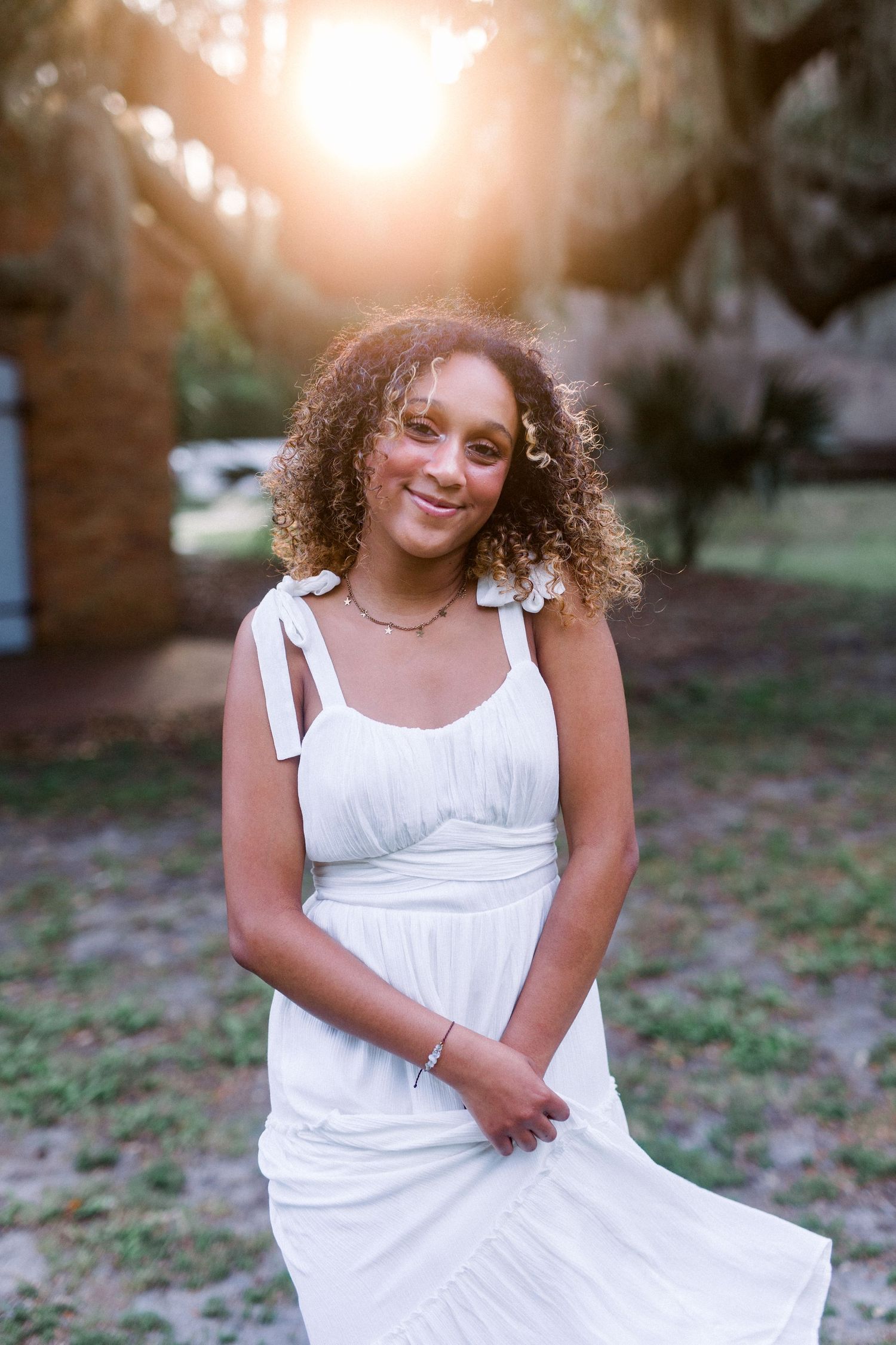 Woman in white tie-shoulder dress poses in garden setting during golden hour.
