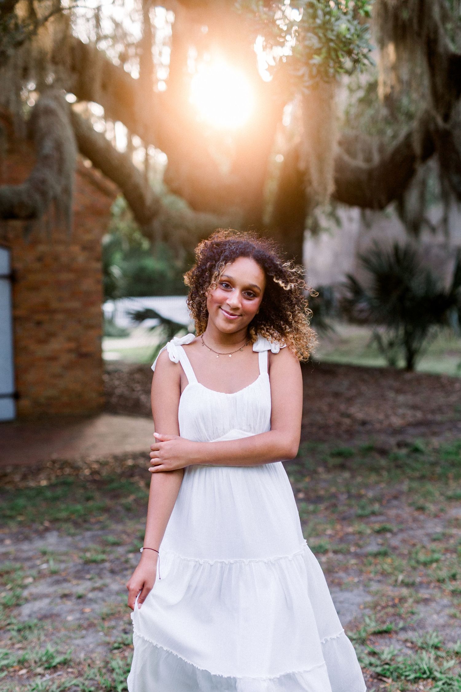 A white sundress photoshoot with natural sunlight peeking through moss-draped trees in a park setting.