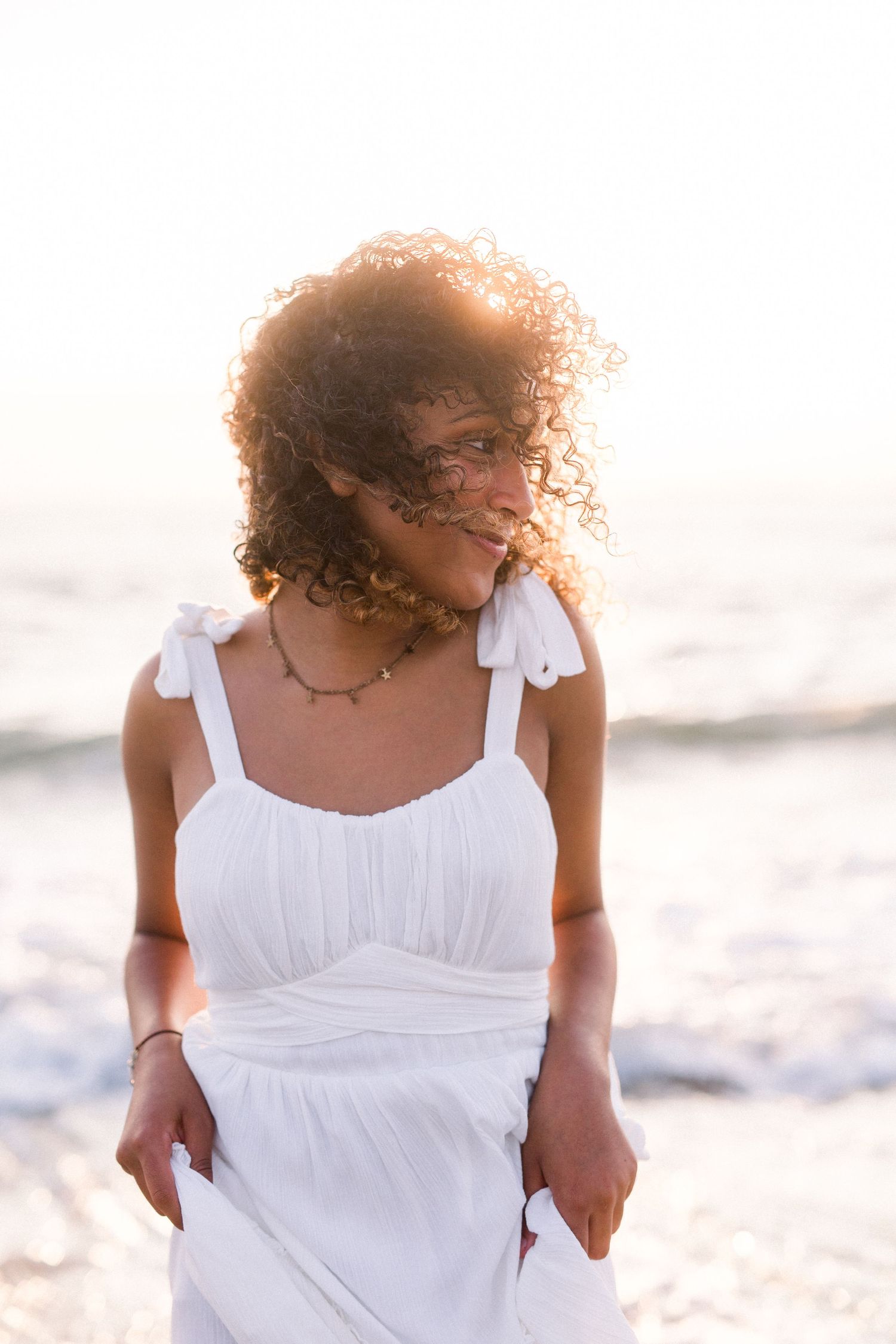 Sunlit beach portrait featuring natural curls and a white summer dress against ocean backdrop.