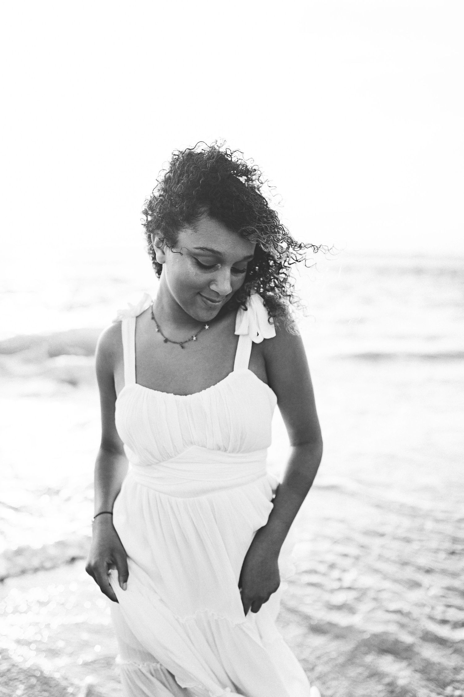 Black and white candid beach photo capturing movement in white dress and curly hair.